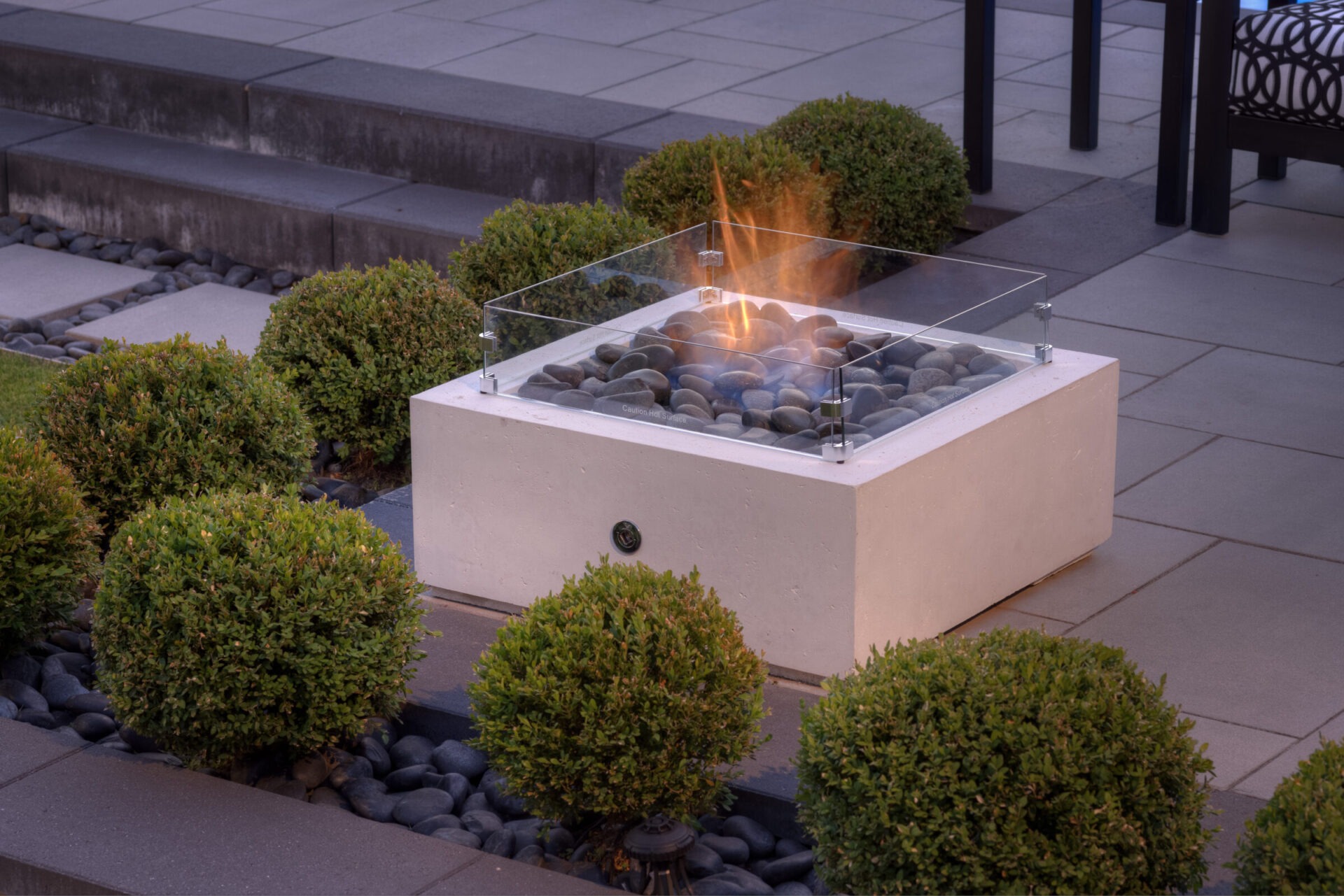 An outdoor gas fire pit with a glass shield surrounded by round bushes and smooth stones, set against a cozy patio with furniture.