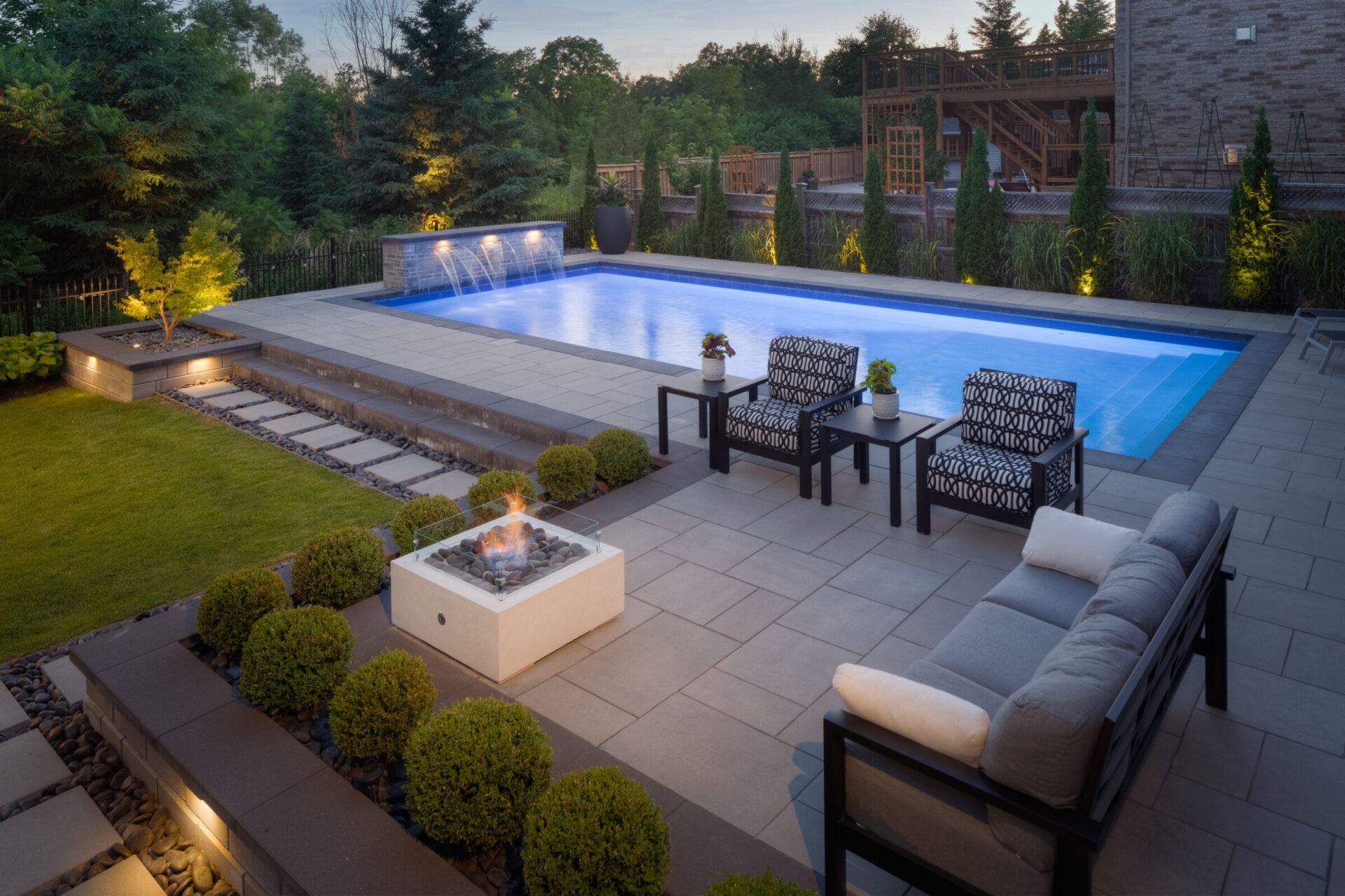 This image shows a luxury backyard at dusk with a lit swimming pool, stylish outdoor furniture, a fire pit, landscaping, and a wooden deck.