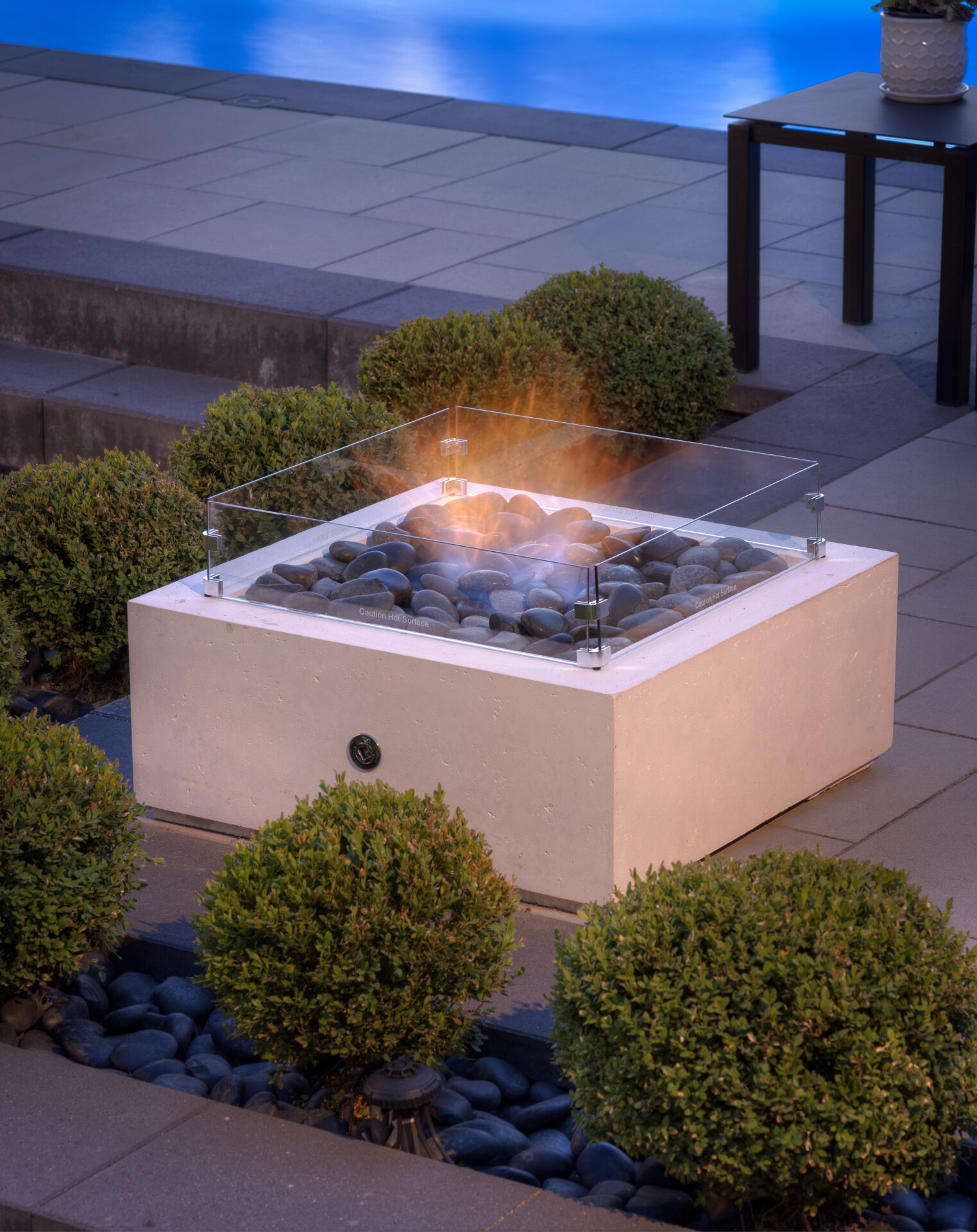 An outdoor gas fire pit with glass wind guard surrounded by dark stones and green shrubbery, adjacent to a patio table, at dusk.