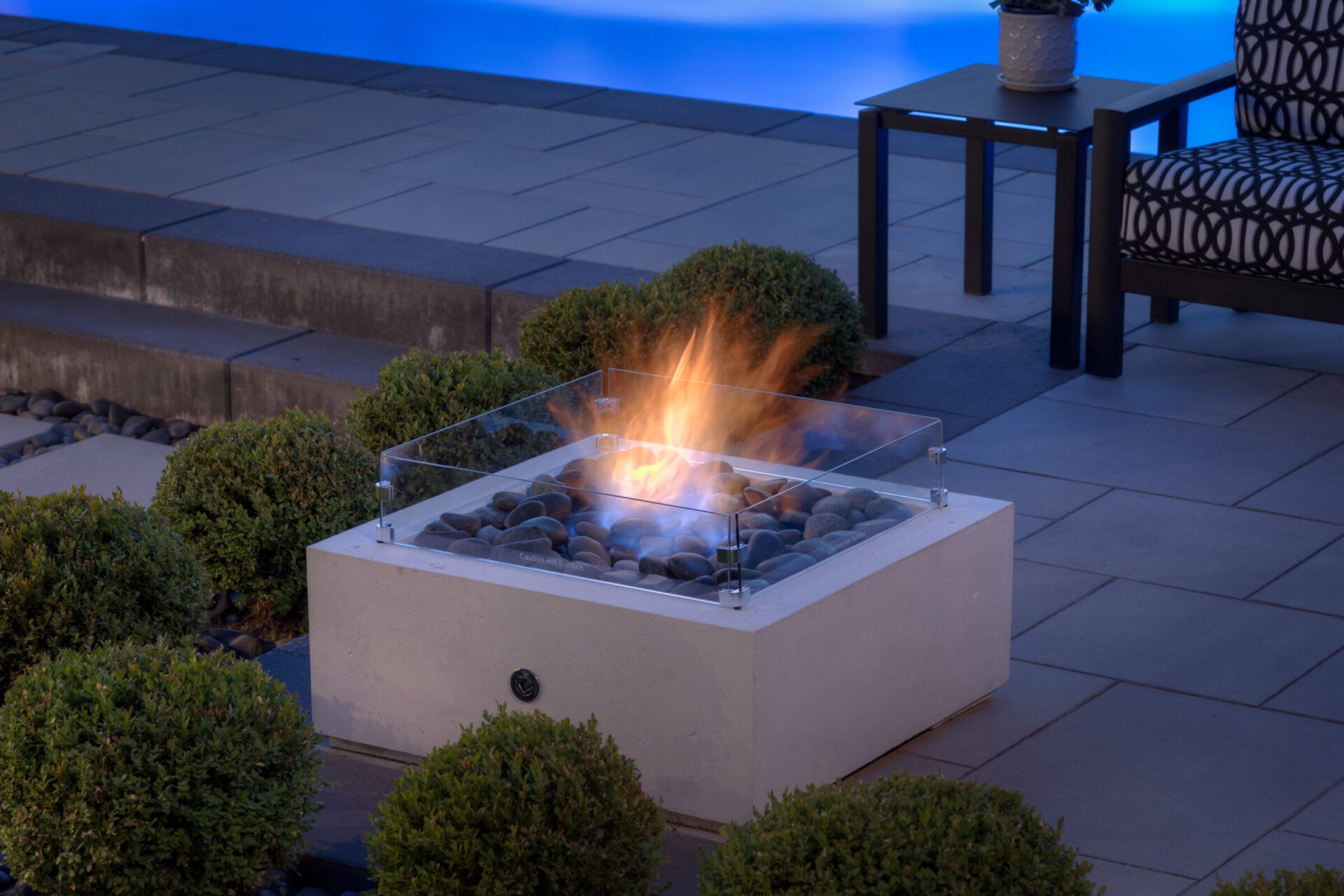 An outdoor glass-enclosed fire pit with a lively flame burns atop smooth stones. Nearby are bushes, a side table, and a patterned armchair. Evening ambiance.