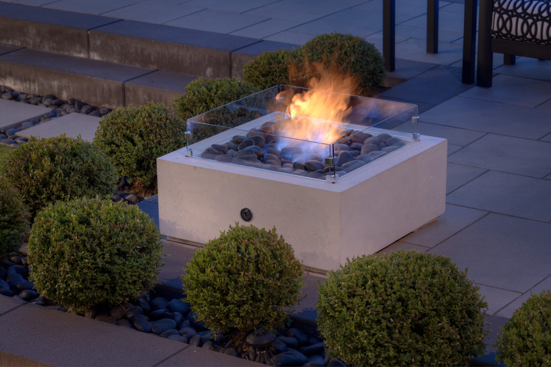 An outdoor patio with a modern square fire pit, featuring clear glass wind guards, surrounded by dark pebbles, green shrubs, and paving stones.