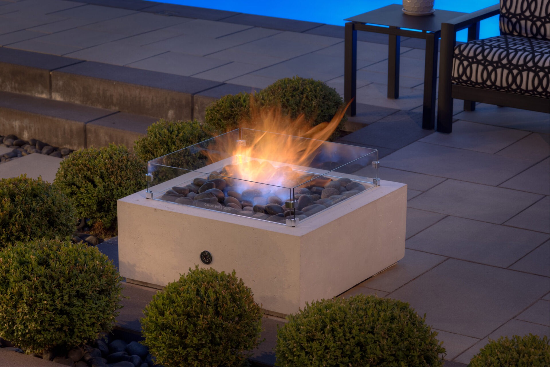 An outdoor gas fire pit with a glass shield surrounded by smooth stones and bushes is shown during twilight, complementing a stylish patio setup.