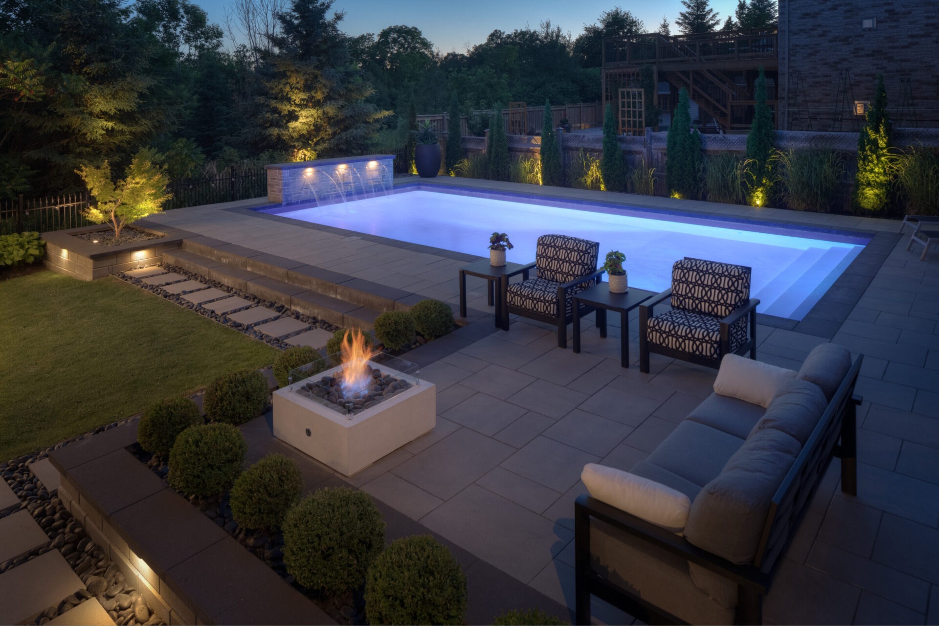 An elegant outdoor setting at dusk featuring a serene swimming pool, comfortable patio furniture, a fire feature, manicured lawn, and ambient lighting.
