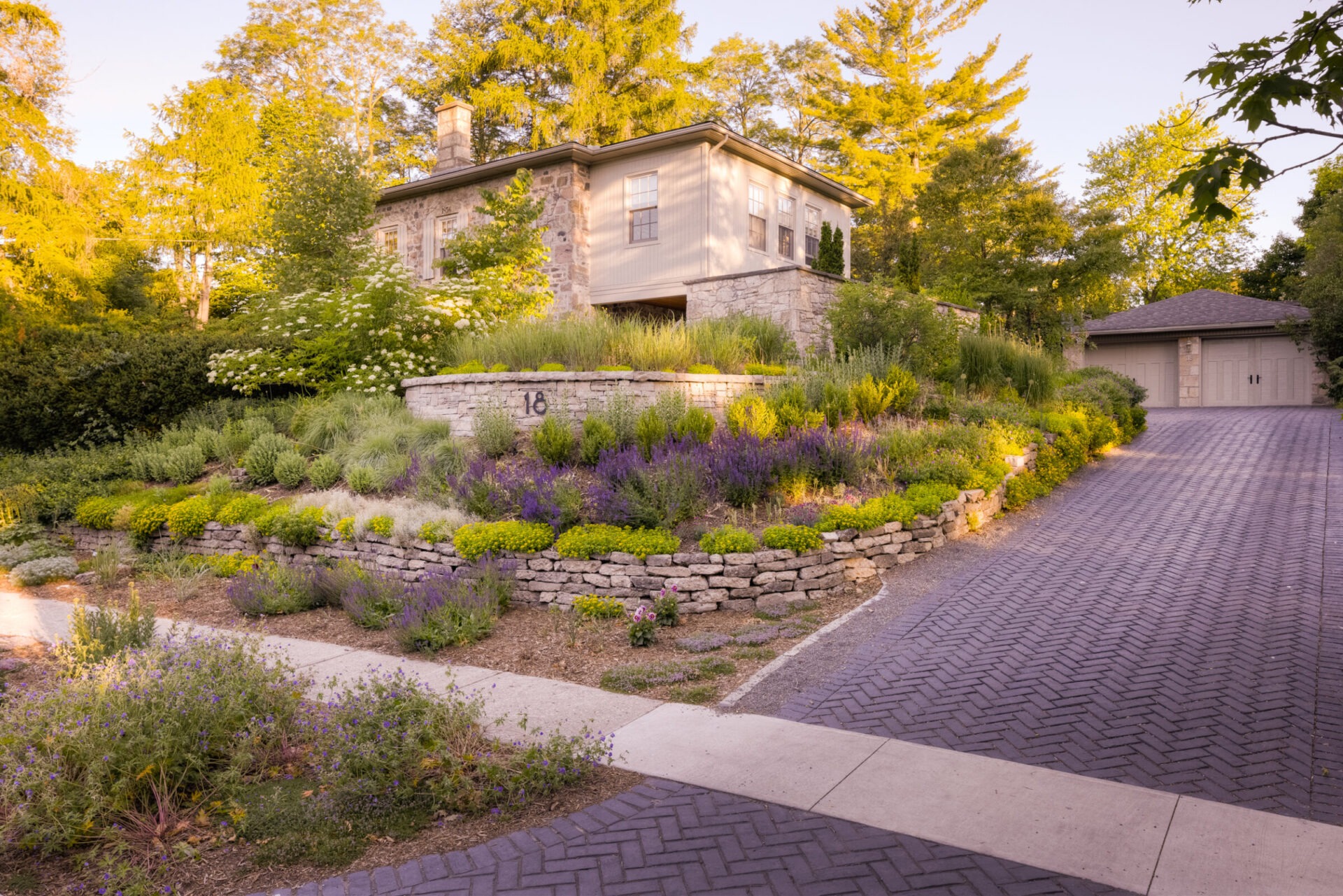 A serene residential setting featuring a stone house with siding, landscaped tiered garden beds, and a cobblestone driveway at golden hour.