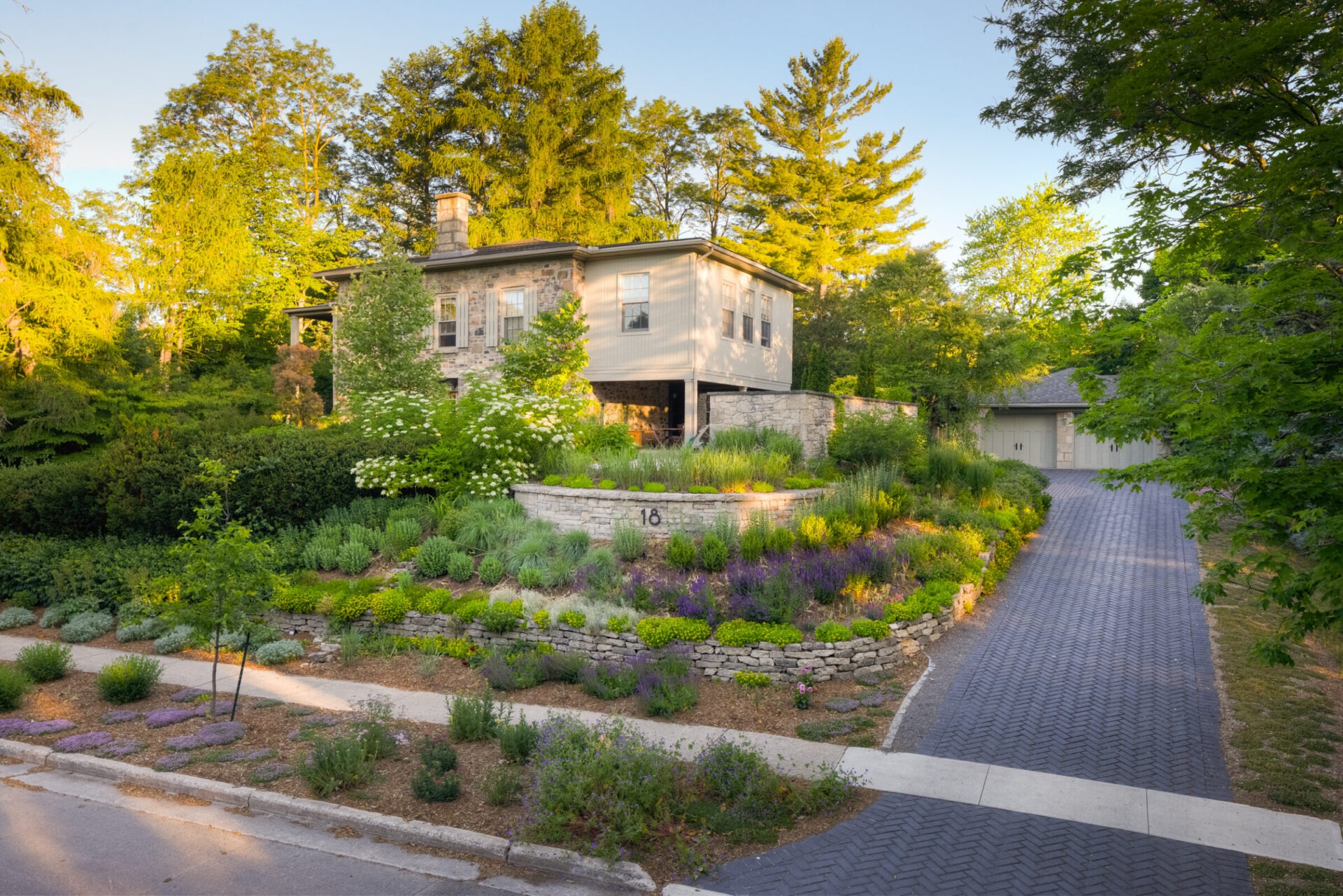 A two-story house with mixed siding, surrounded by lush landscaping and mature trees, sits beside a cobblestone driveway during golden hour.