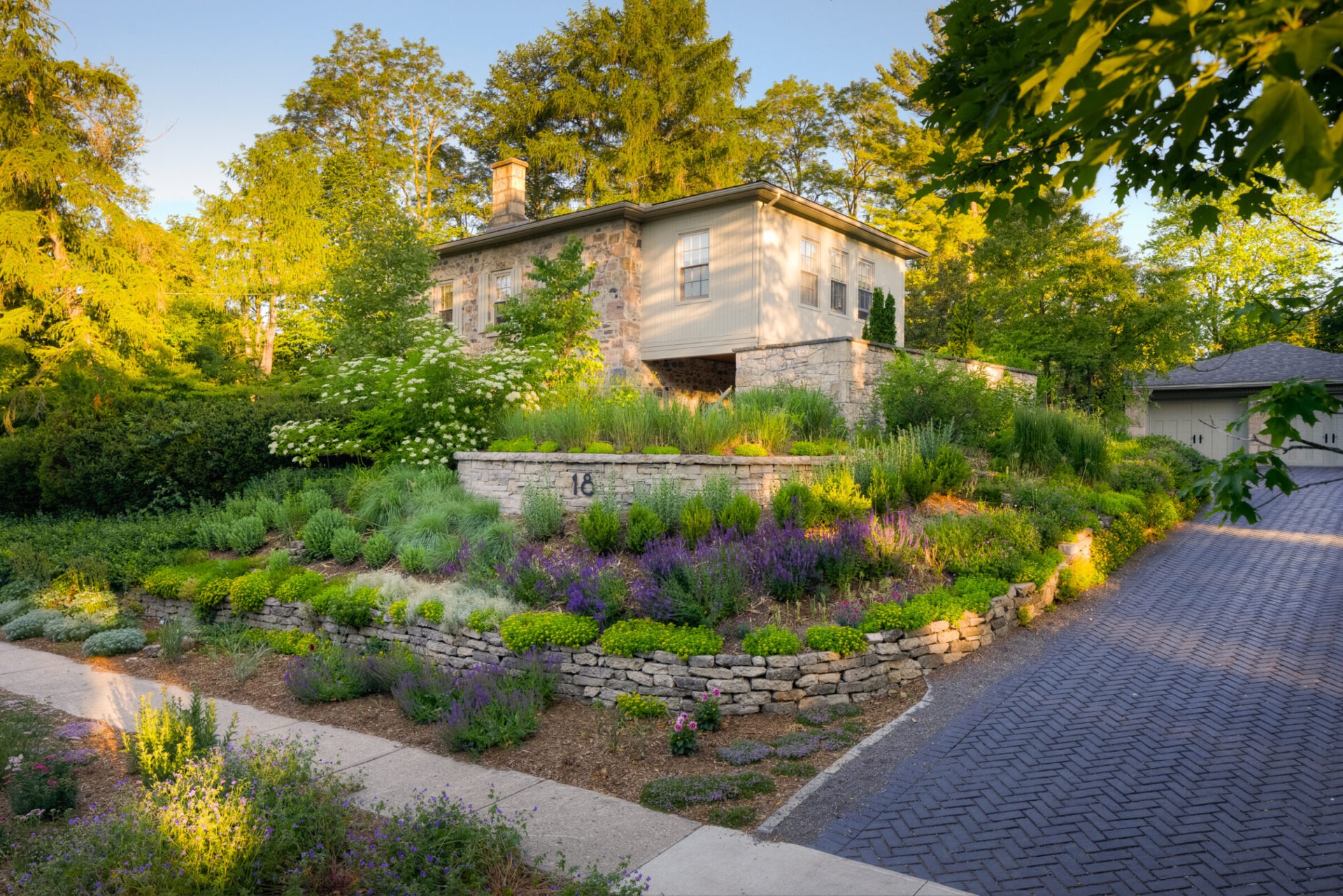 A stone house with beige siding sits amidst lush landscaping with blooming purple flowers, greenery, and a cobblestone driveway during sunset.
