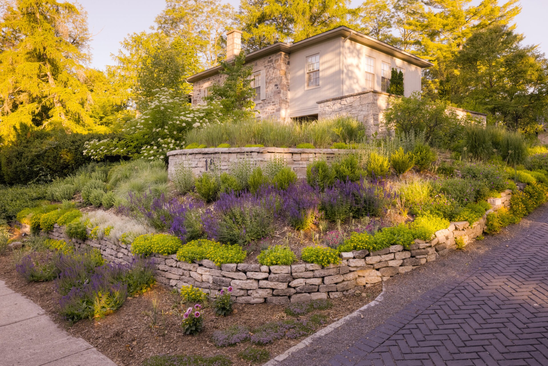 A quaint stone house overlooks a beautifully landscaped garden with lavender, green shrubs, and a cobblestone path during a warm, golden sunset.