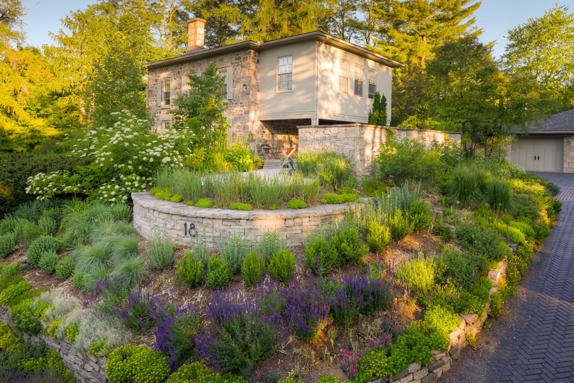A stone house with a modern extension surrounded by lush, tiered landscaping under a warm sunset light. Cobblestone driveway leads to a garage.