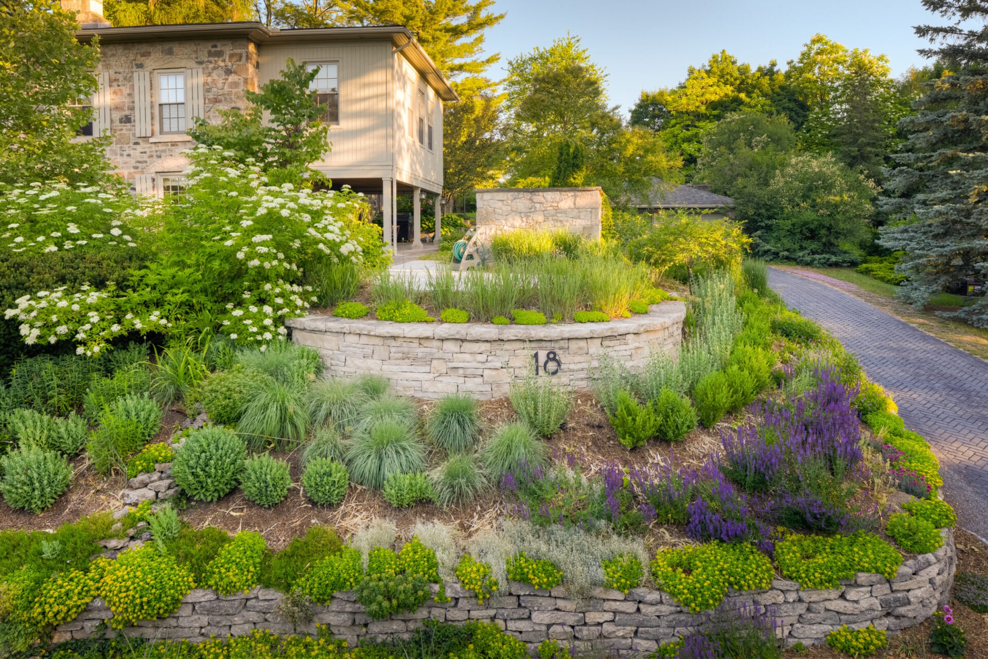 This image showcases a landscaped garden, with tiered stone walls, flourishing greenery, and vibrant flowers, in front of a traditional stone-faced house.