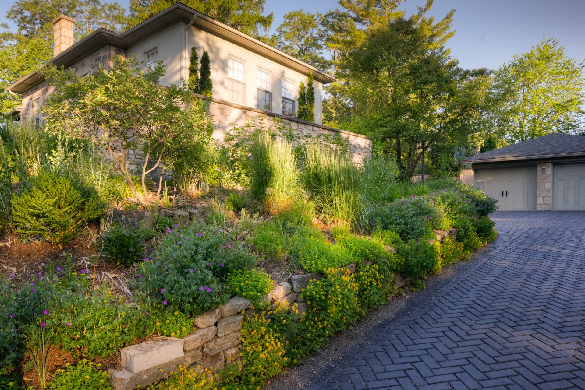 A well-landscaped property with lush gardens and a cobblestone driveway leading to a detached garage, adjacent to a house with stone accents.
