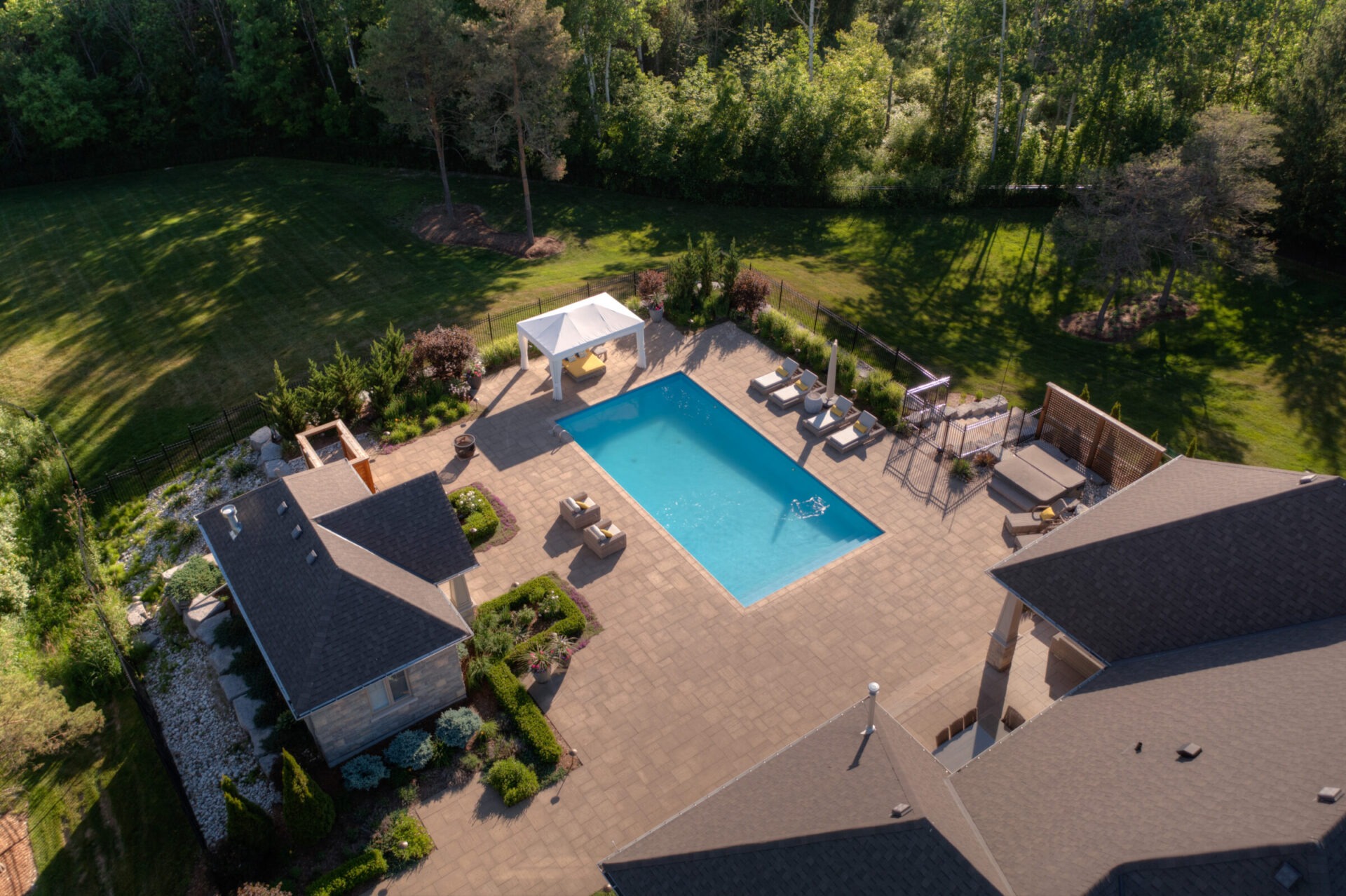Aerial view of a residential backyard with an inground swimming pool, patio area, outdoor furniture, neatly landscaped garden, and adjoining house roofs.