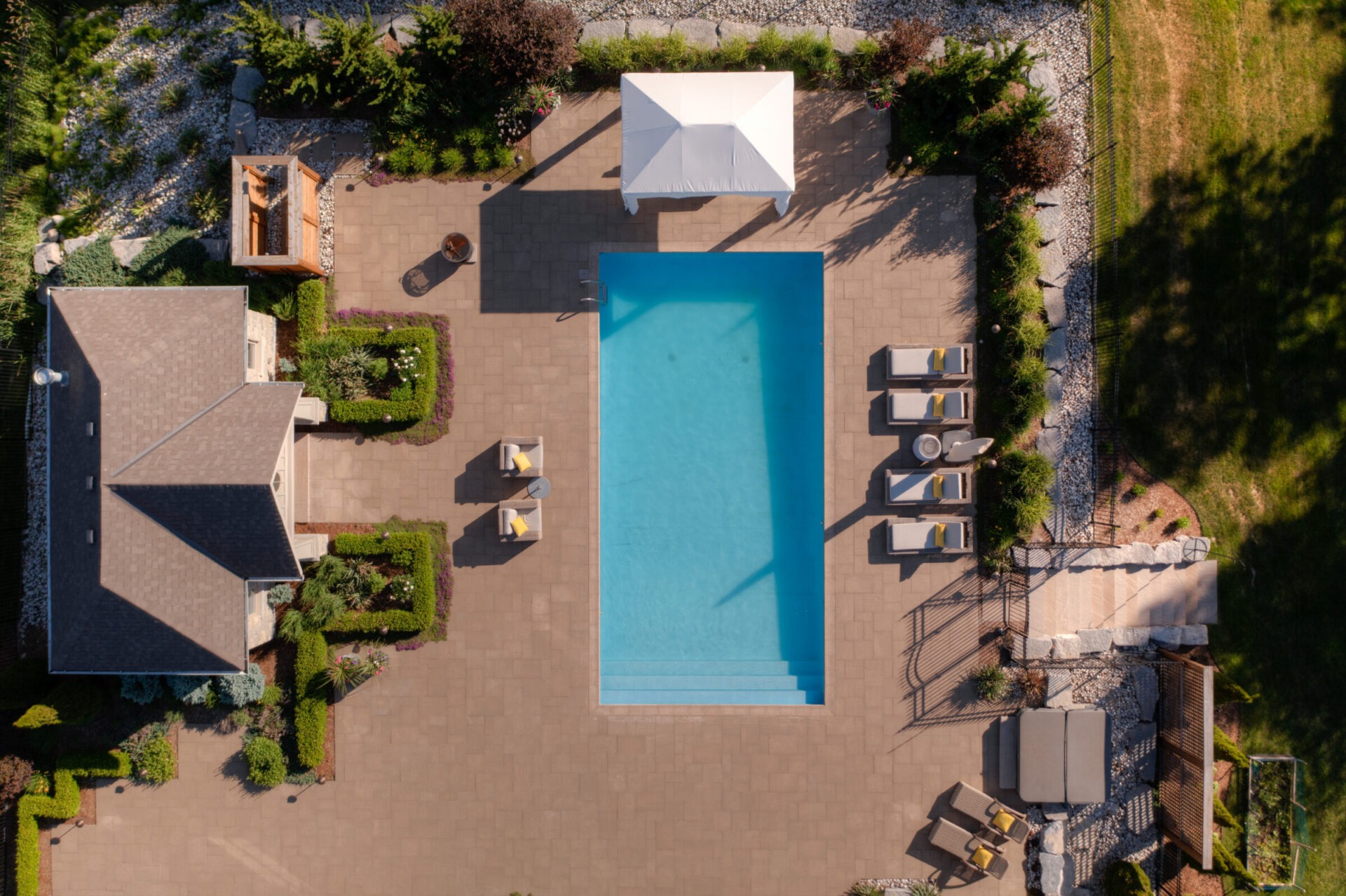 Aerial view of a neat backyard with a large rectangular pool, patio, loungers, landscaped garden, and sections of a residential building.
