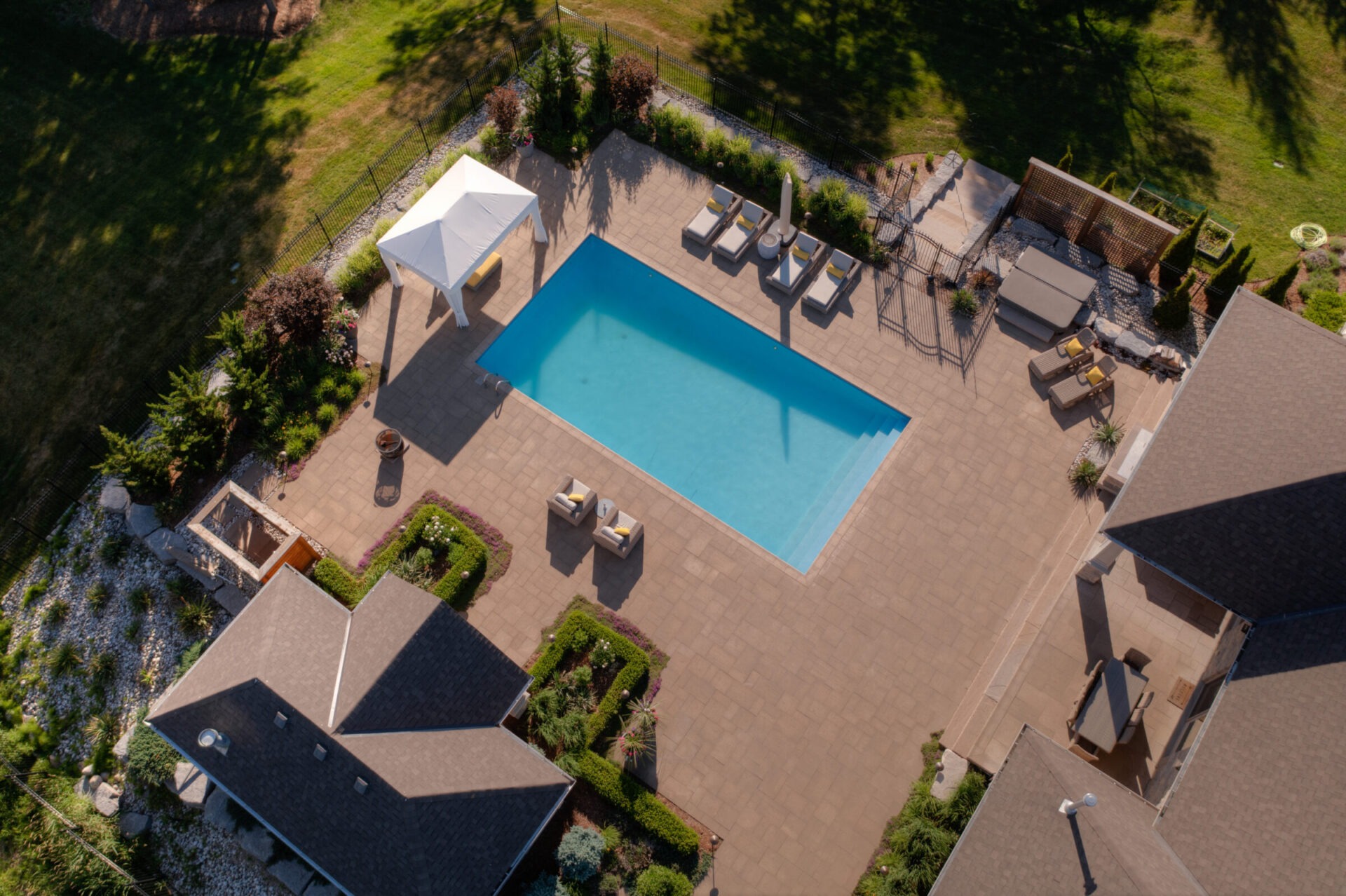 Aerial view of a residential backyard with a large swimming pool, patio area, sun loungers, landscaping, a gazebo, and part of two houses.