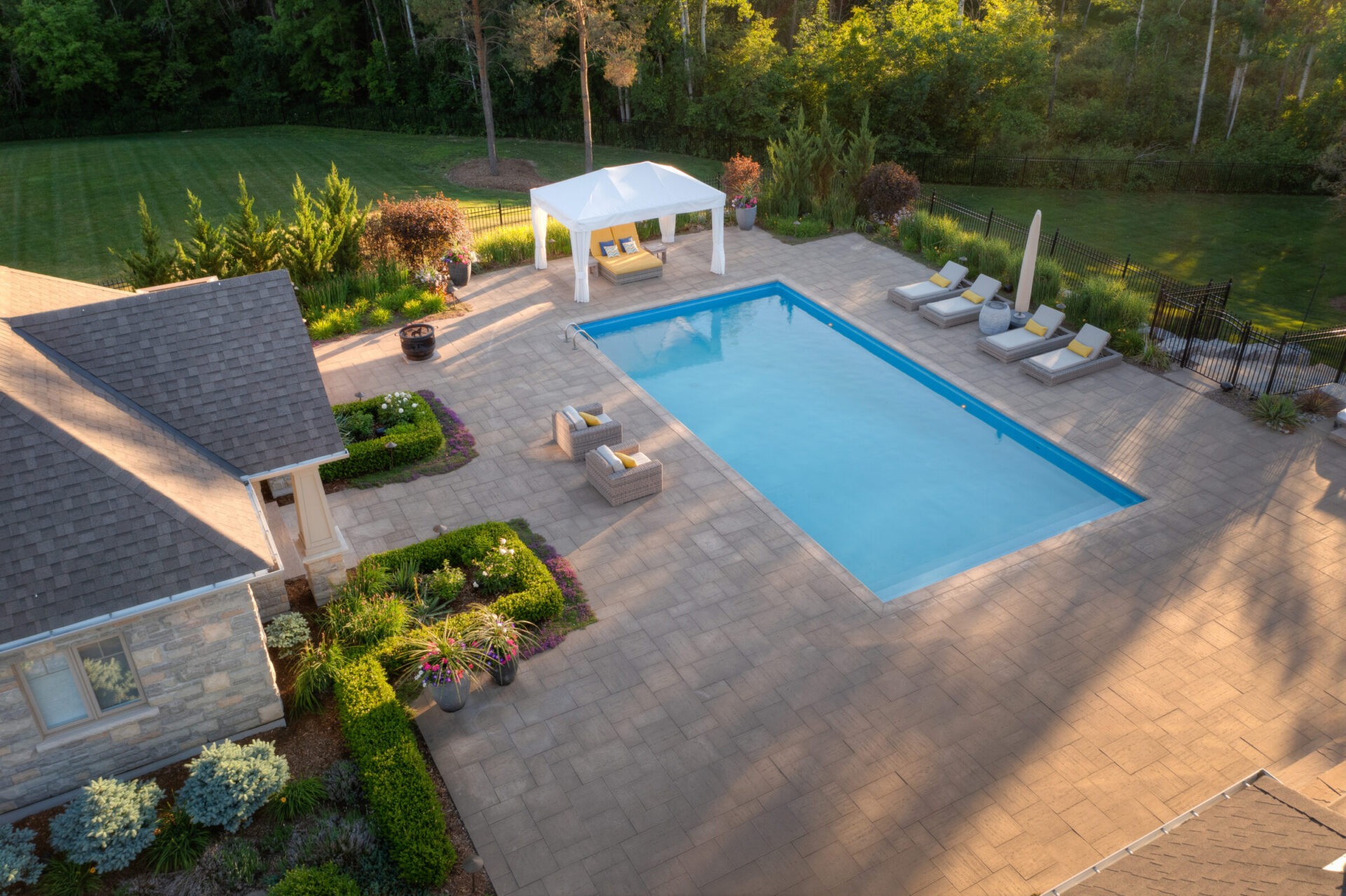 An aerial view of a backyard with a rectangular pool, patio area, sun loungers, manicured garden, and a canopy, bordered by trees at dusk.