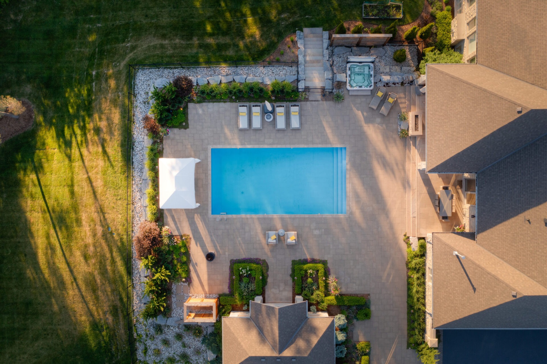 Aerial view of a residential backyard with a rectangular pool, lounge chairs, jacuzzi, manicured gardens, and a house during the golden hour.