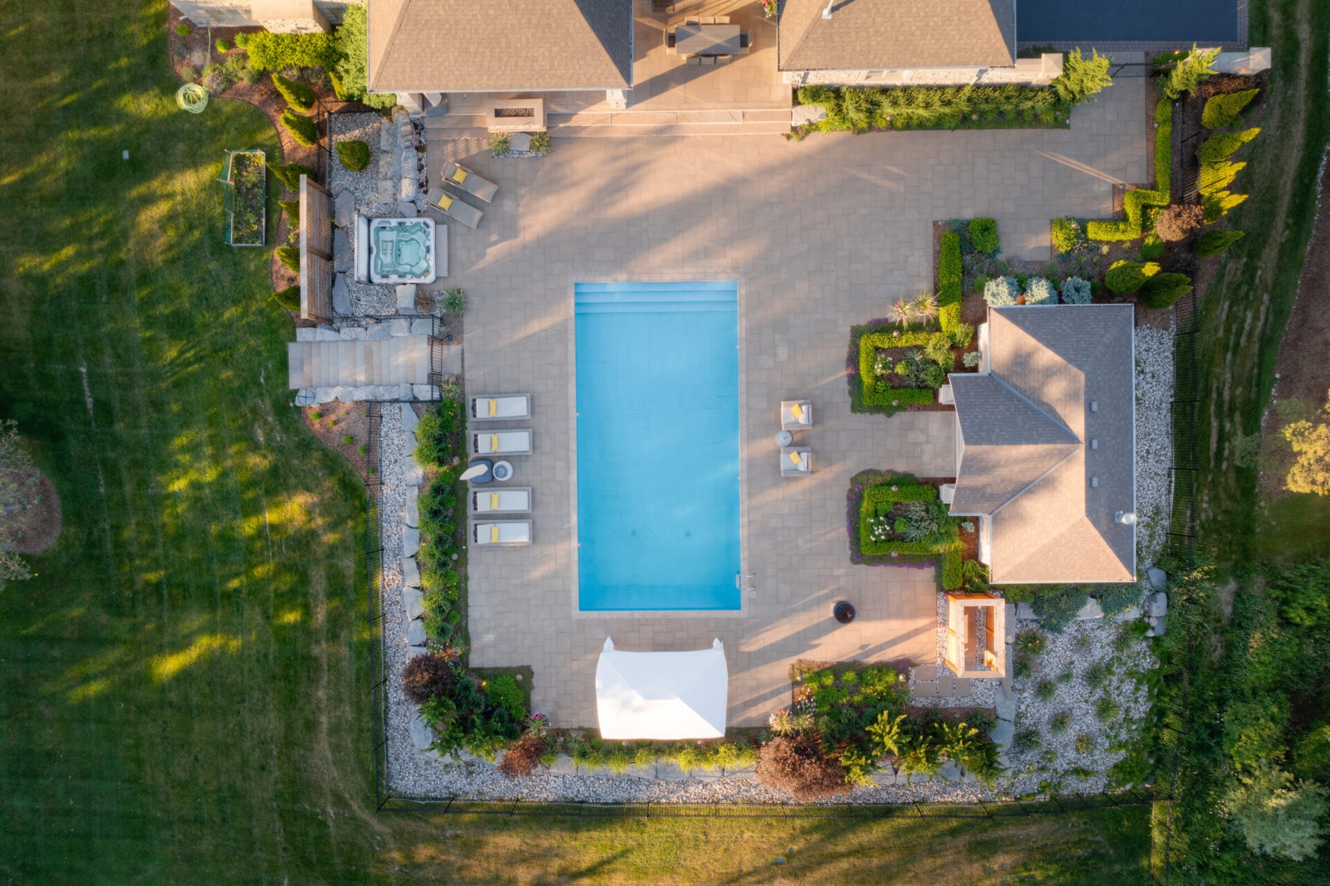 Aerial view of a luxury backyard with a large swimming pool, hot tub, landscaped garden, sun loungers, and a patio area during sunset.