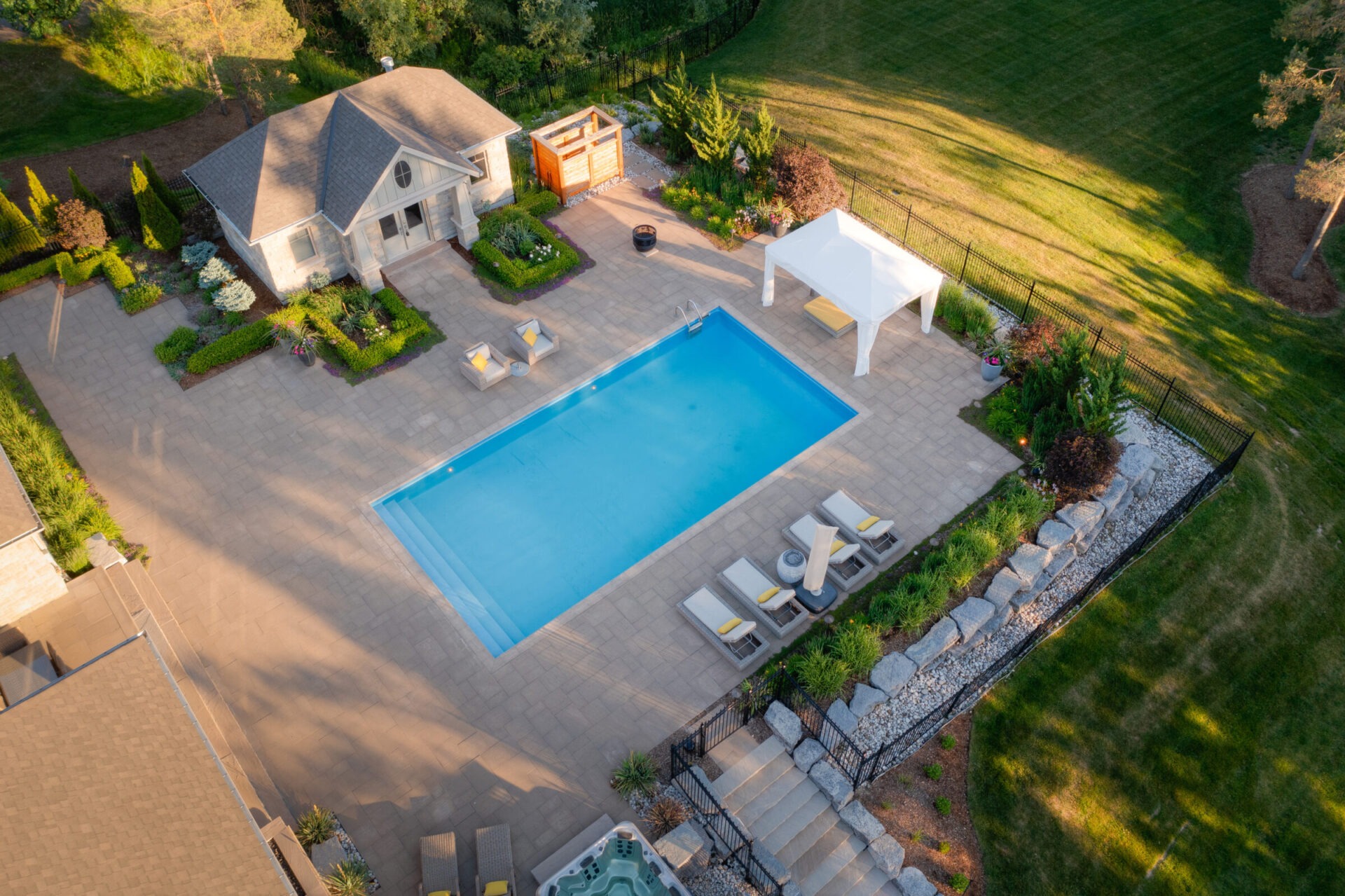 Aerial view of a backyard with a large swimming pool, patio, sun loungers, a gazebo, landscaping, and a hot tub adjoining a residential house.