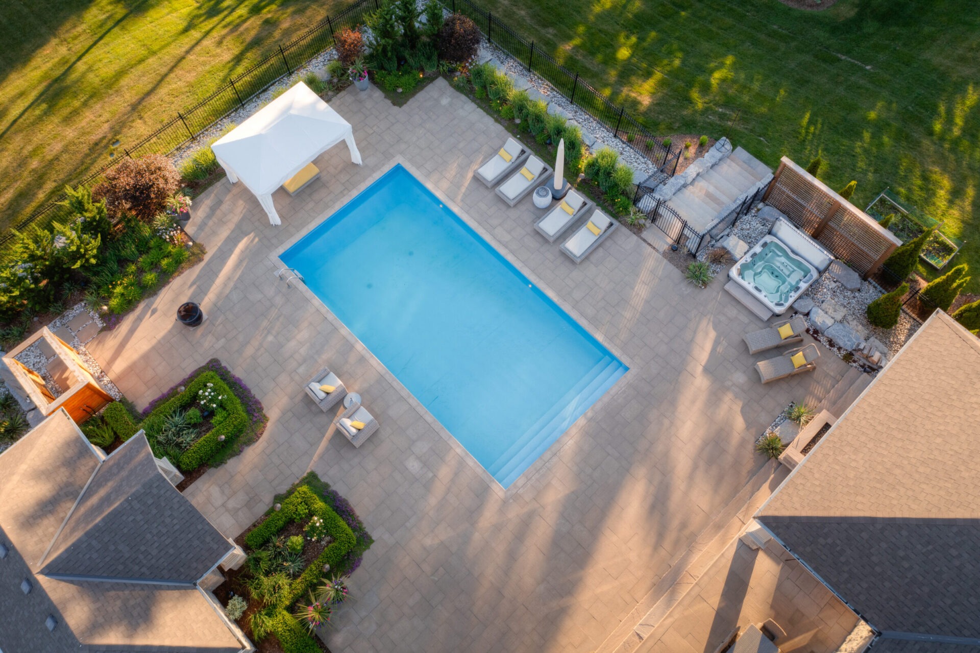 Aerial view of a residential backyard with a rectangular swimming pool, hot tub, lounge chairs, gazebo, manicured lawn, and fenced perimeter at dusk.