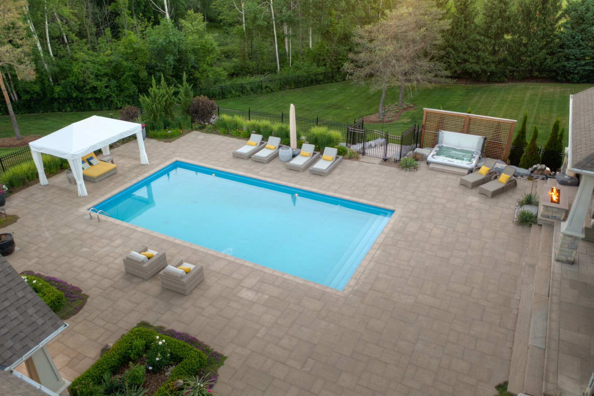 An aerial view of a backyard with a rectangular swimming pool, lounge chairs, a jacuzzi, a pergola, fire pit, and landscaped garden near a forest.