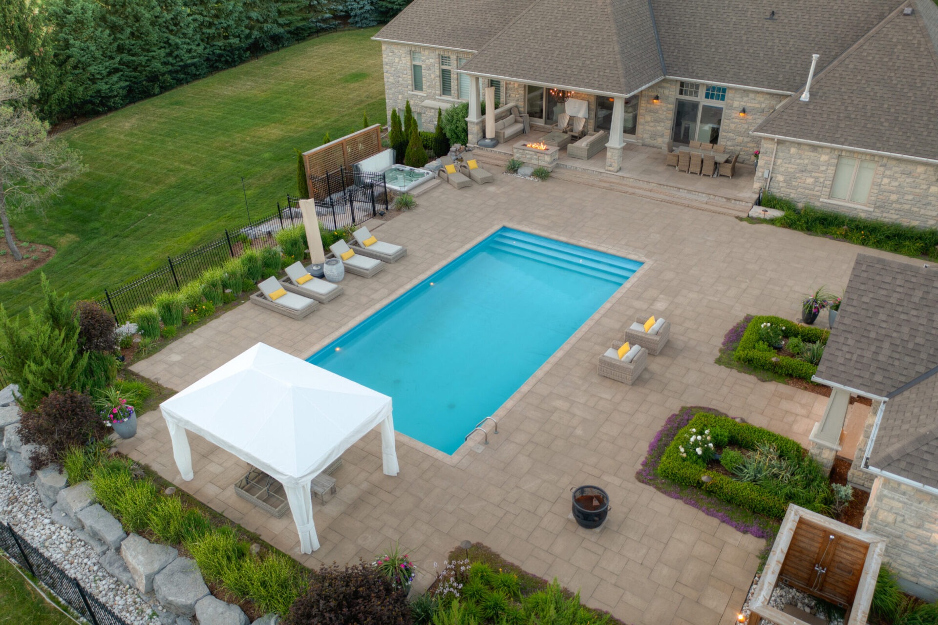 This is an aerial view of a backyard with a rectangular pool, patio, outdoor furniture, a gazebo, a hot tub, and landscaped garden areas.