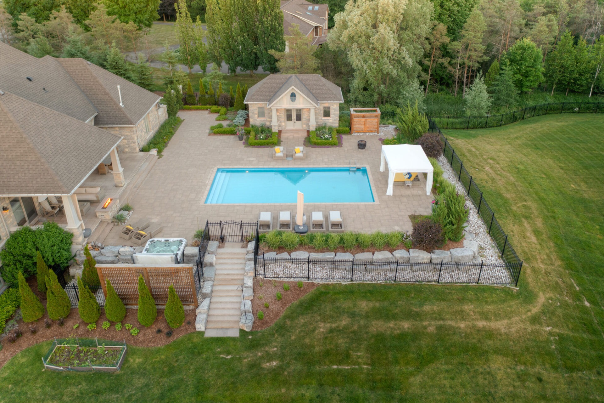 Aerial view of a luxurious backyard with a large pool, hot tub, manicured lawn, patio area, and a pool house surrounded by trees and shrubbery.