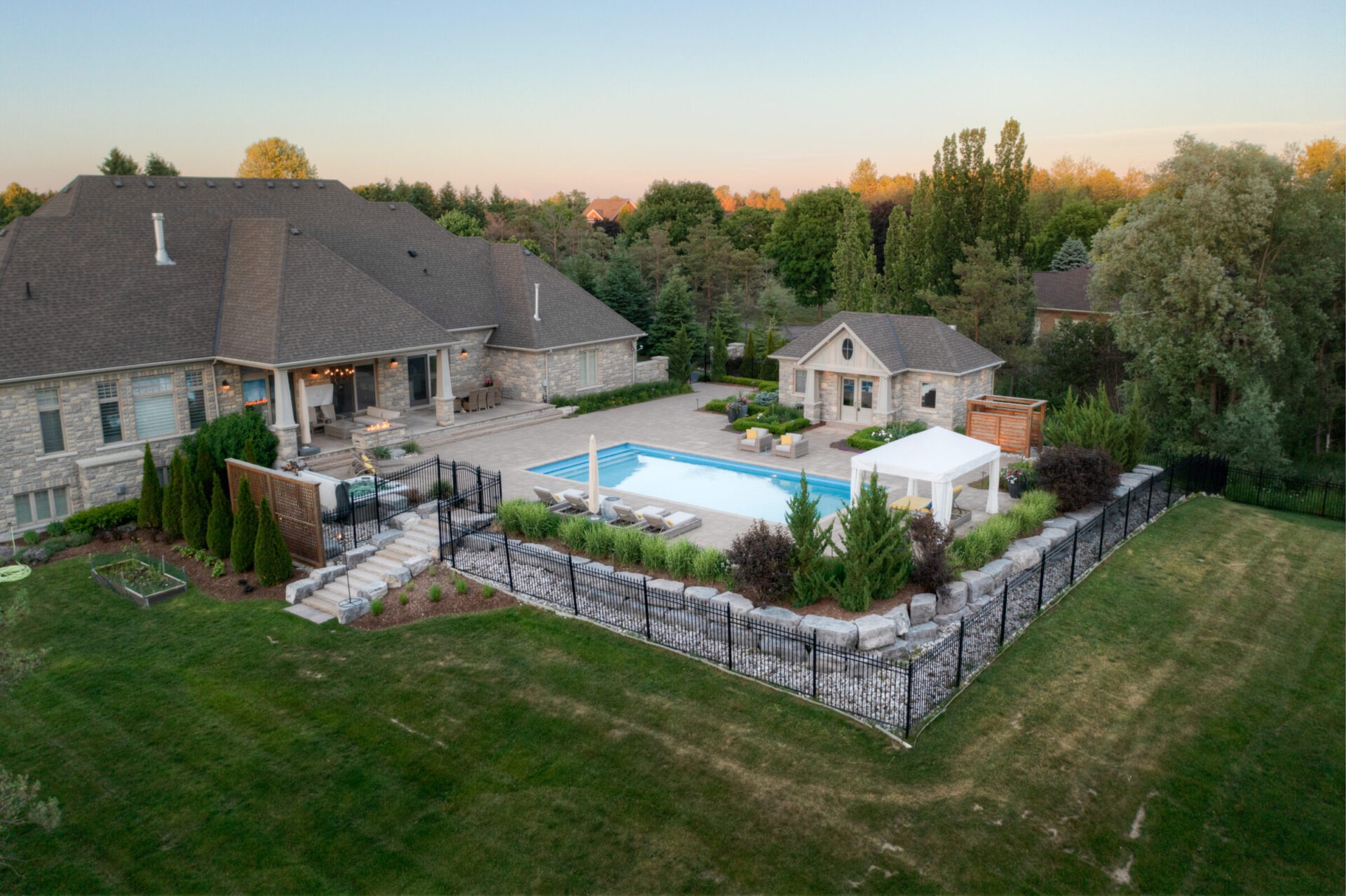 This image features a large, luxurious stone house with a swimming pool, a pool house, and a well-manicured lawn during twilight.