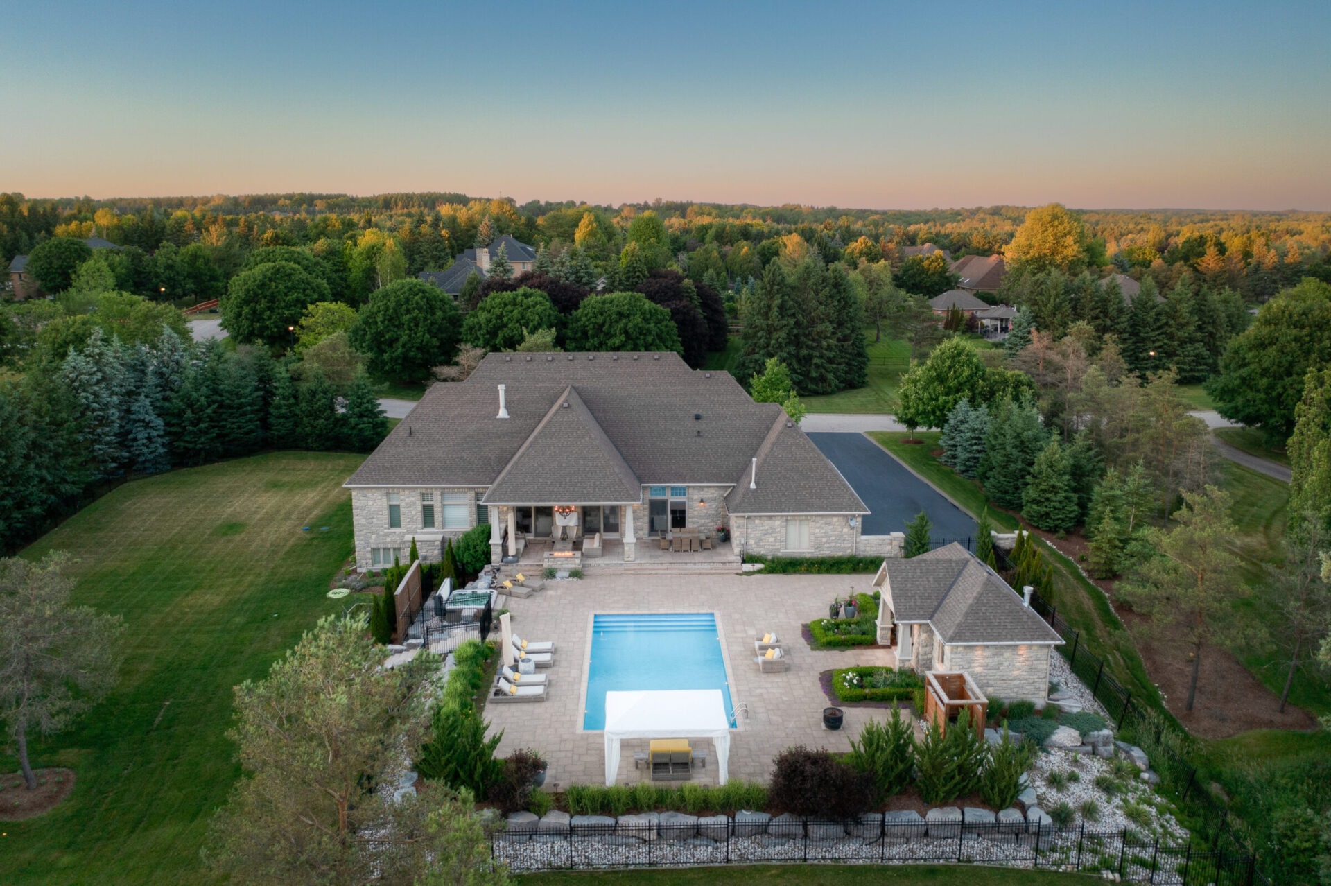 An aerial view shows a luxurious house with a large swimming pool, landscaped garden, and surrounding trees during twilight. No people are visible.