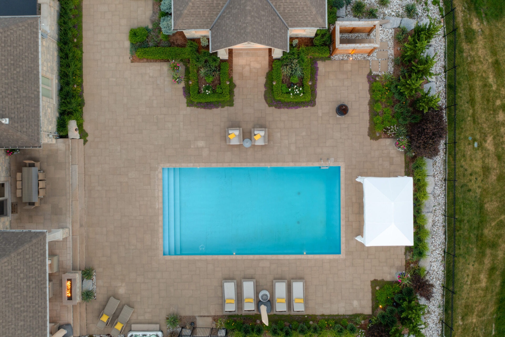 Aerial view of a backyard with a large rectangular swimming pool, paved area, sun loungers, manicured gardens, and a small outdoor structure.