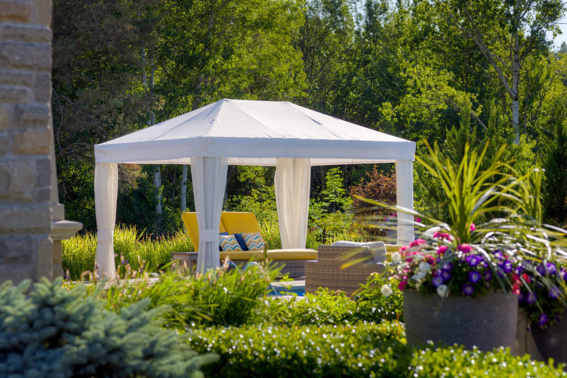 A serene garden setting features a white gazebo with sheer curtains and cozy yellow-cushioned patio furniture surrounded by lush greenery and vibrant flowers.