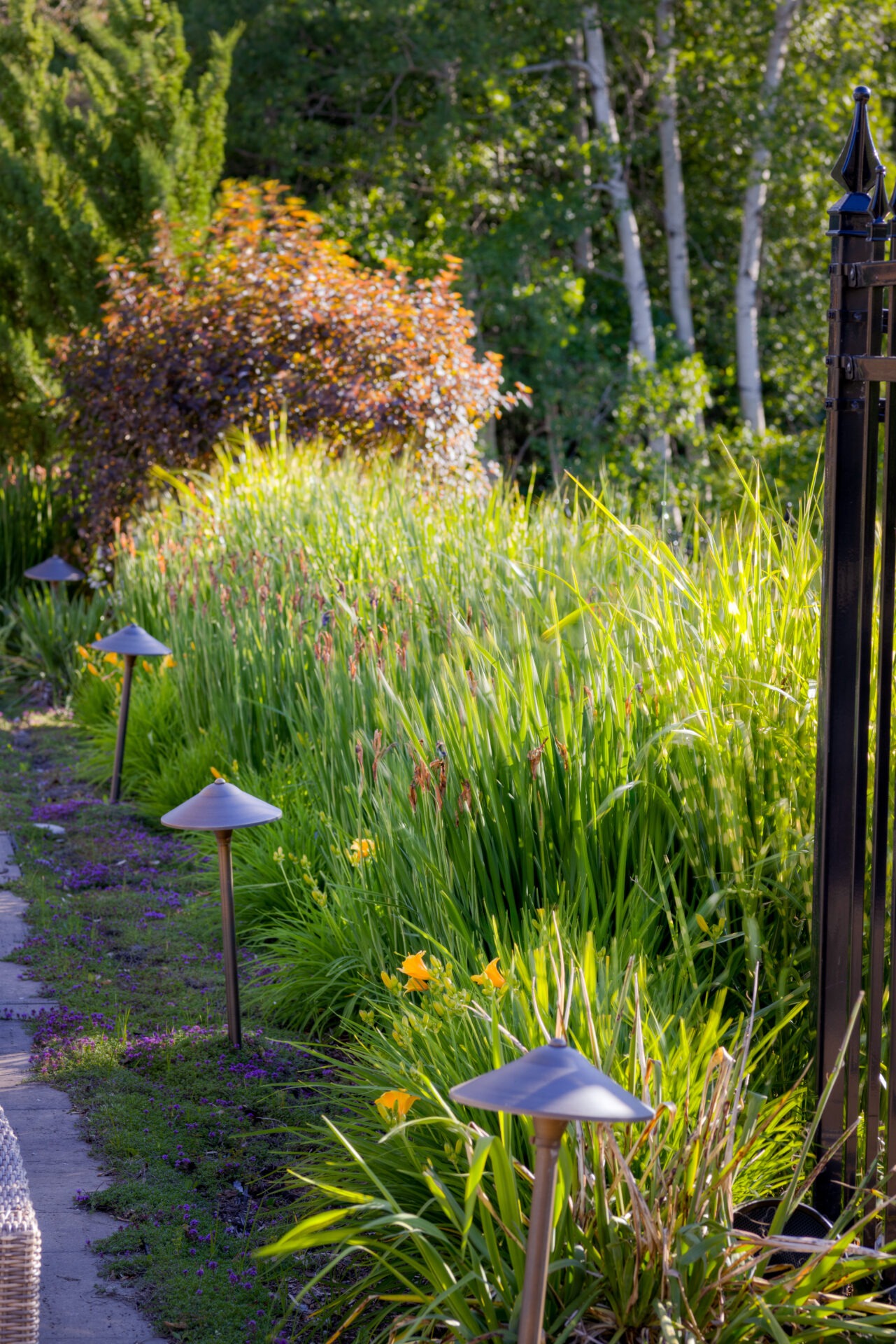 A serene garden path lined with green foliage and yellow flowers, illuminated by mushroom-shaped lights beside a decorative wrought iron fence.