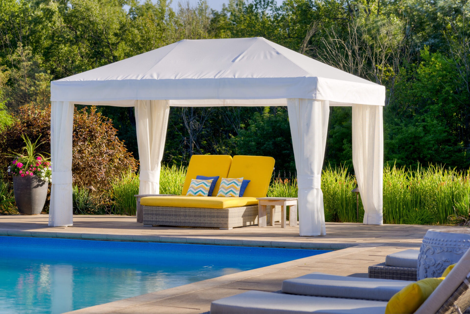 A luxurious poolside features a white gazebo with flowing curtains, a bright yellow lounger with decorative pillows, surrounded by lush greenery and vibrant plants.