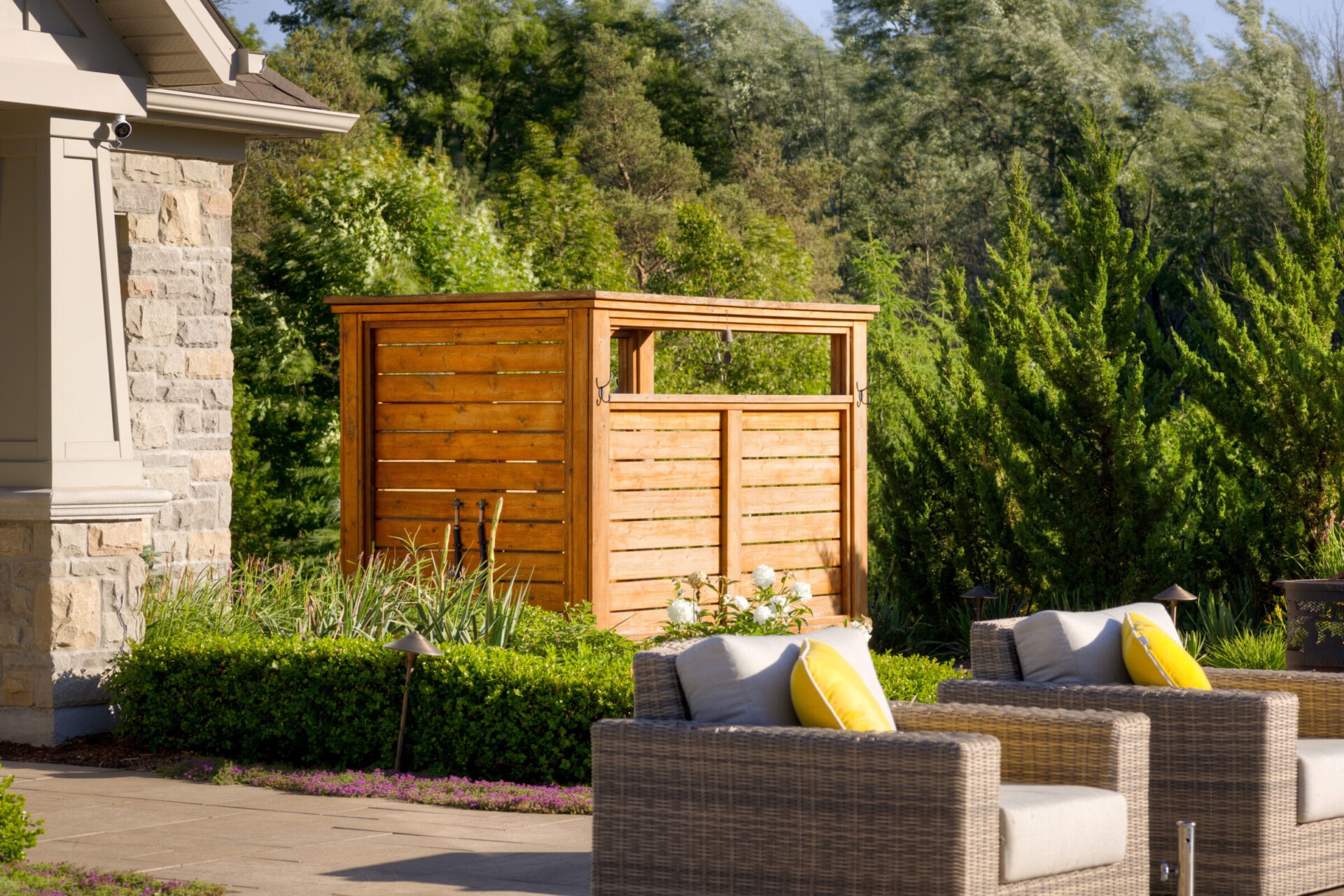 A cozy outdoor patio with wicker furniture and yellow cushions, adjacent to lush greenery and a stylish wooden privacy screen, under clear skies.