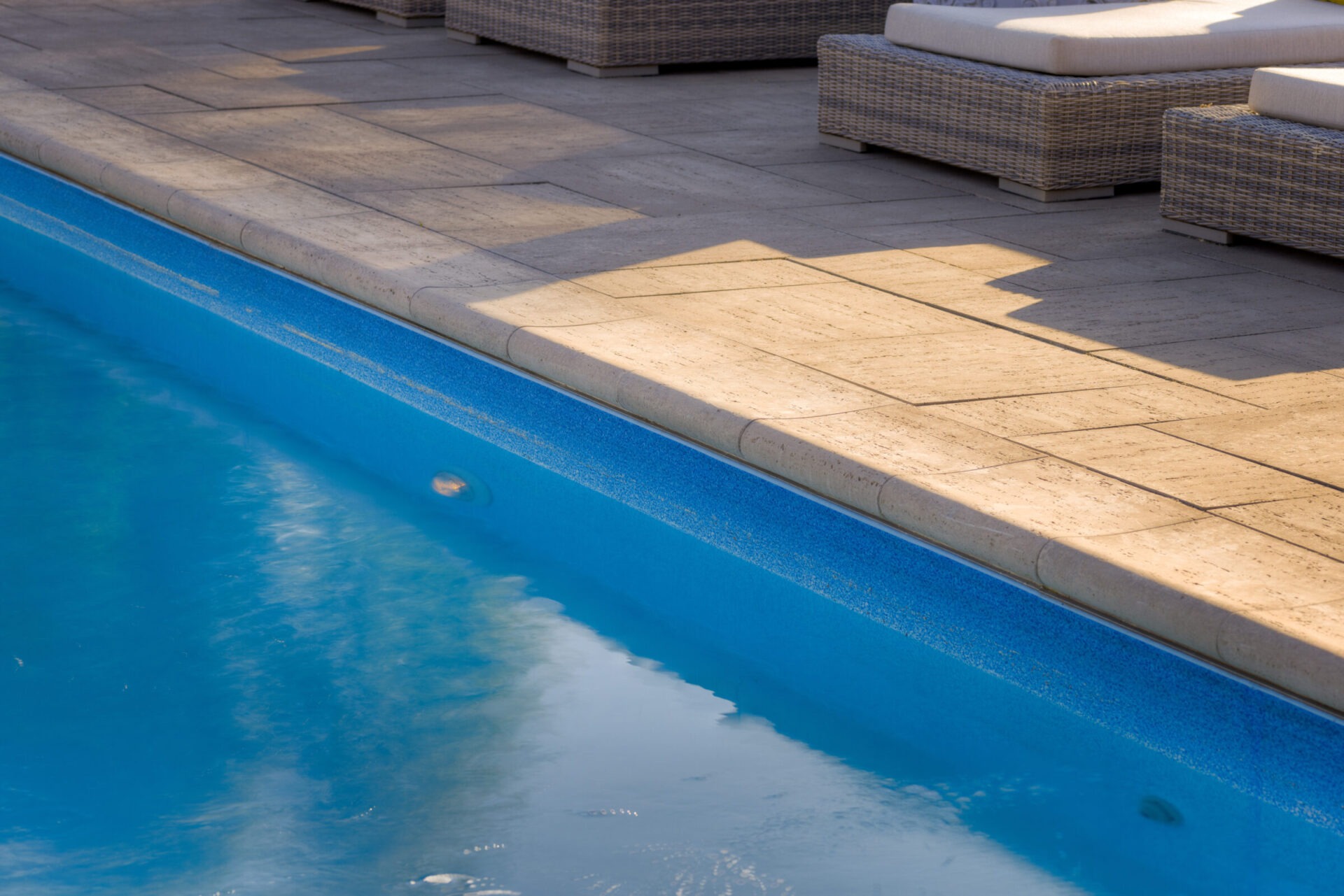 A serene pool with calm blue water adjacent to a beige tiled deck, shaded by lounges, reflecting a clear sky, conveying relaxation and tranquility.