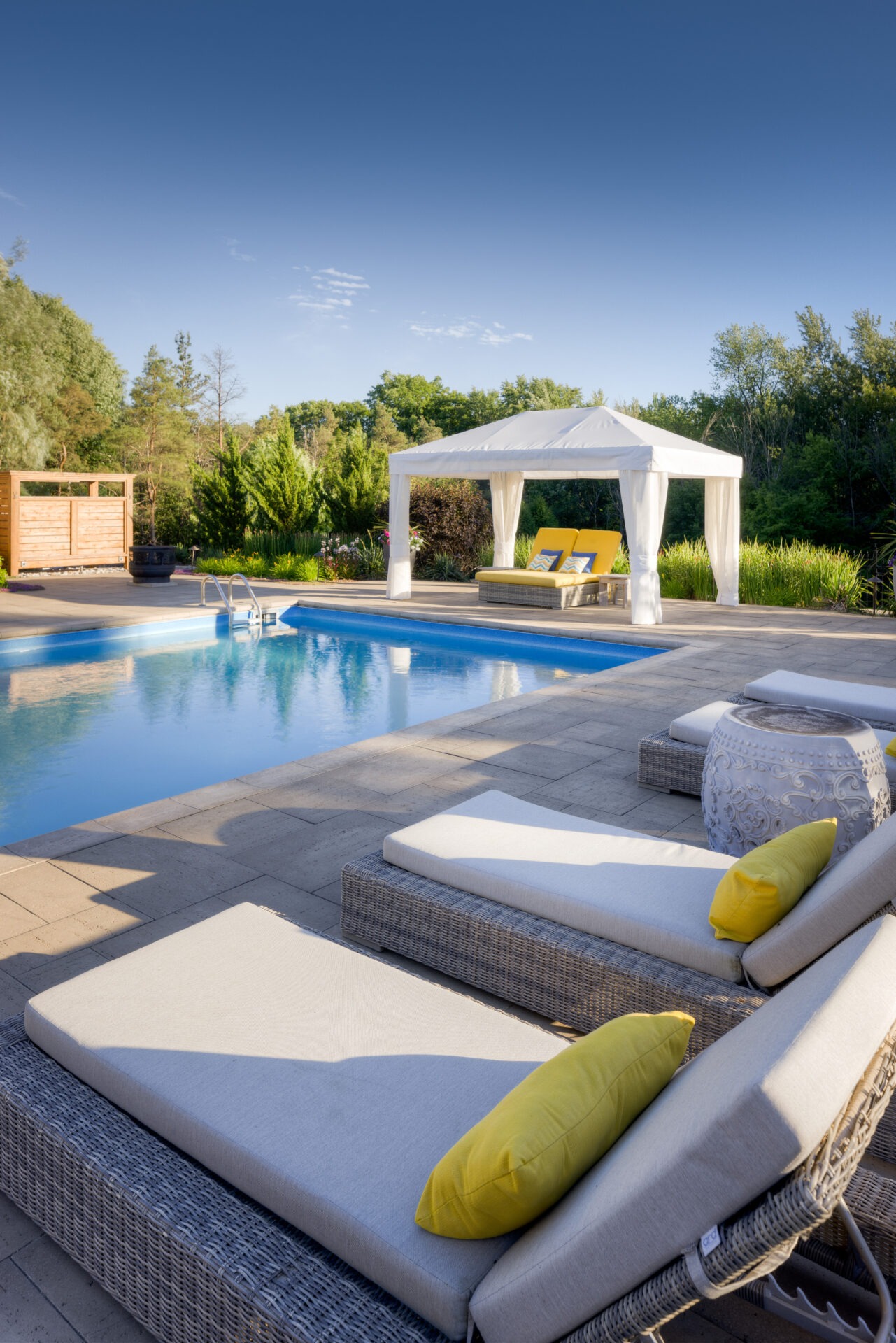 Luxurious outdoor pool surrounded by lounge chairs and a canopy bed, with lush greenery under a clear blue sky, suggesting a tranquil private retreat.