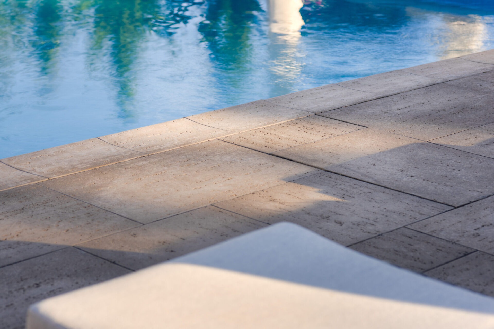 This is a tranquil scene showing the edge of a swimming pool with clear, reflective water, bordered by neatly laid beige tiles and a white ledge.