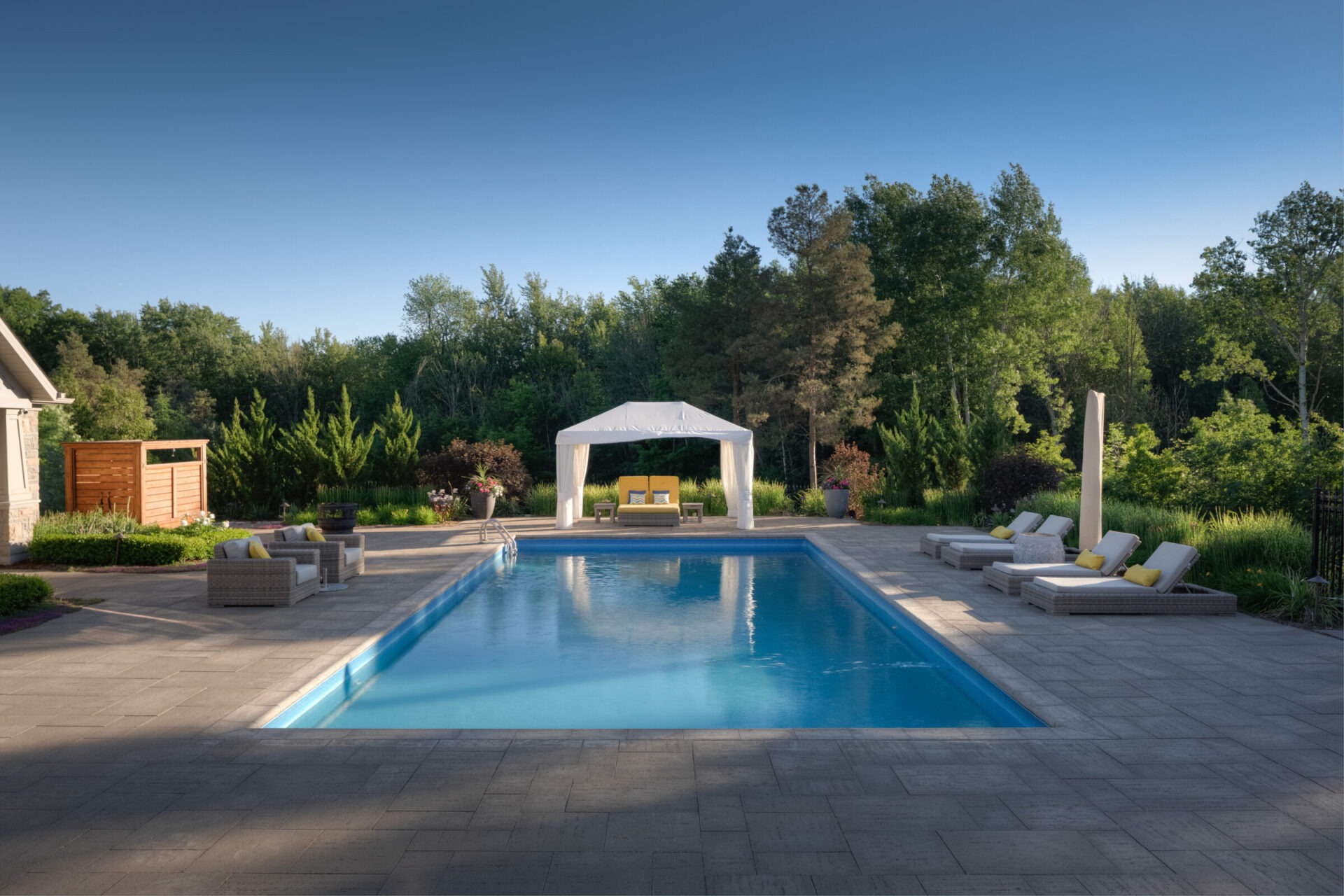 An elegant outdoor setting with a rectangular swimming pool, surrounded by lounge chairs, lush greenery, and a gazebo, under a clear blue sky.