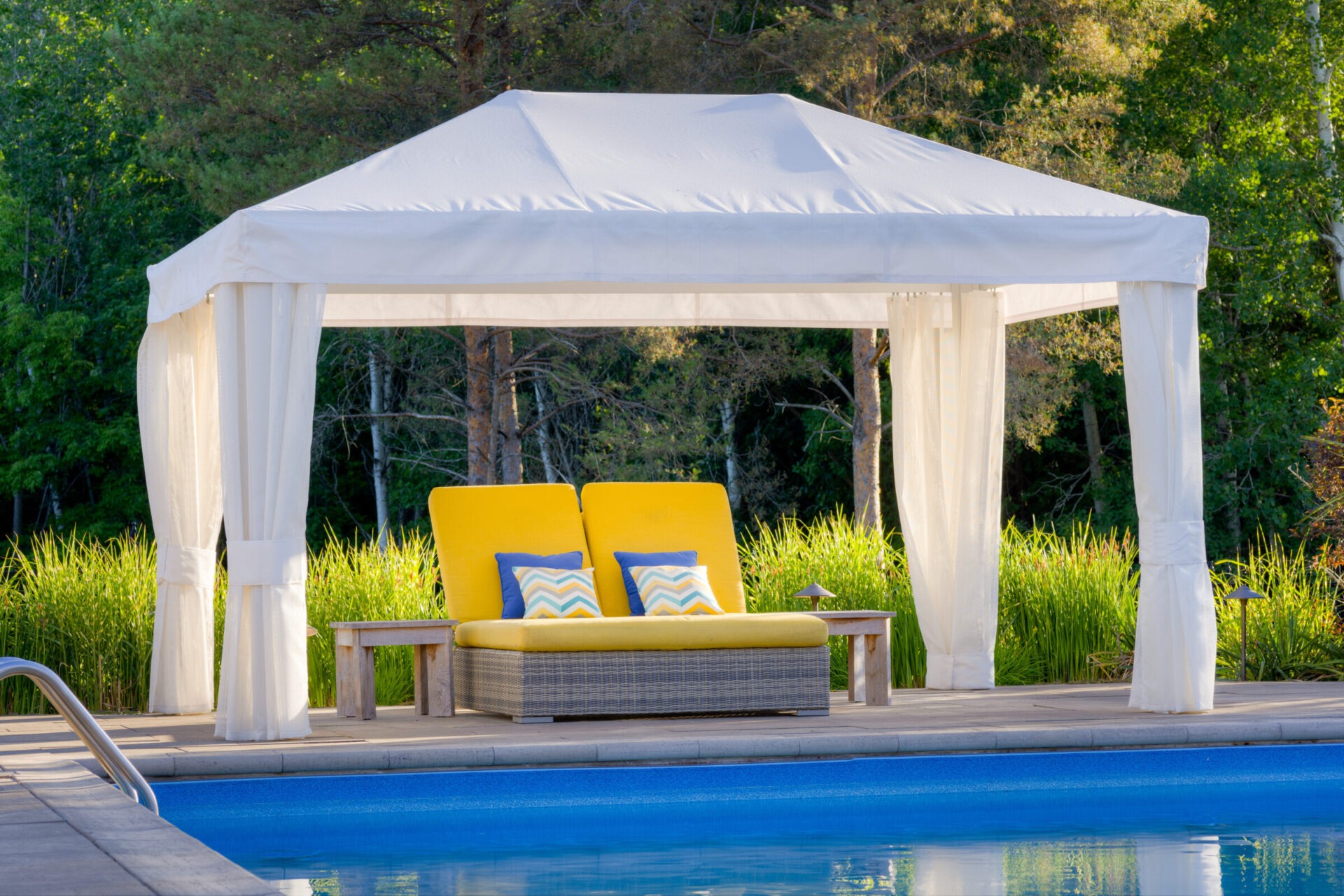 A white poolside gazebo with sheer curtains shelters a wicker loveseat with bright yellow cushions and decorative pillows, beside a tranquil swimming pool.