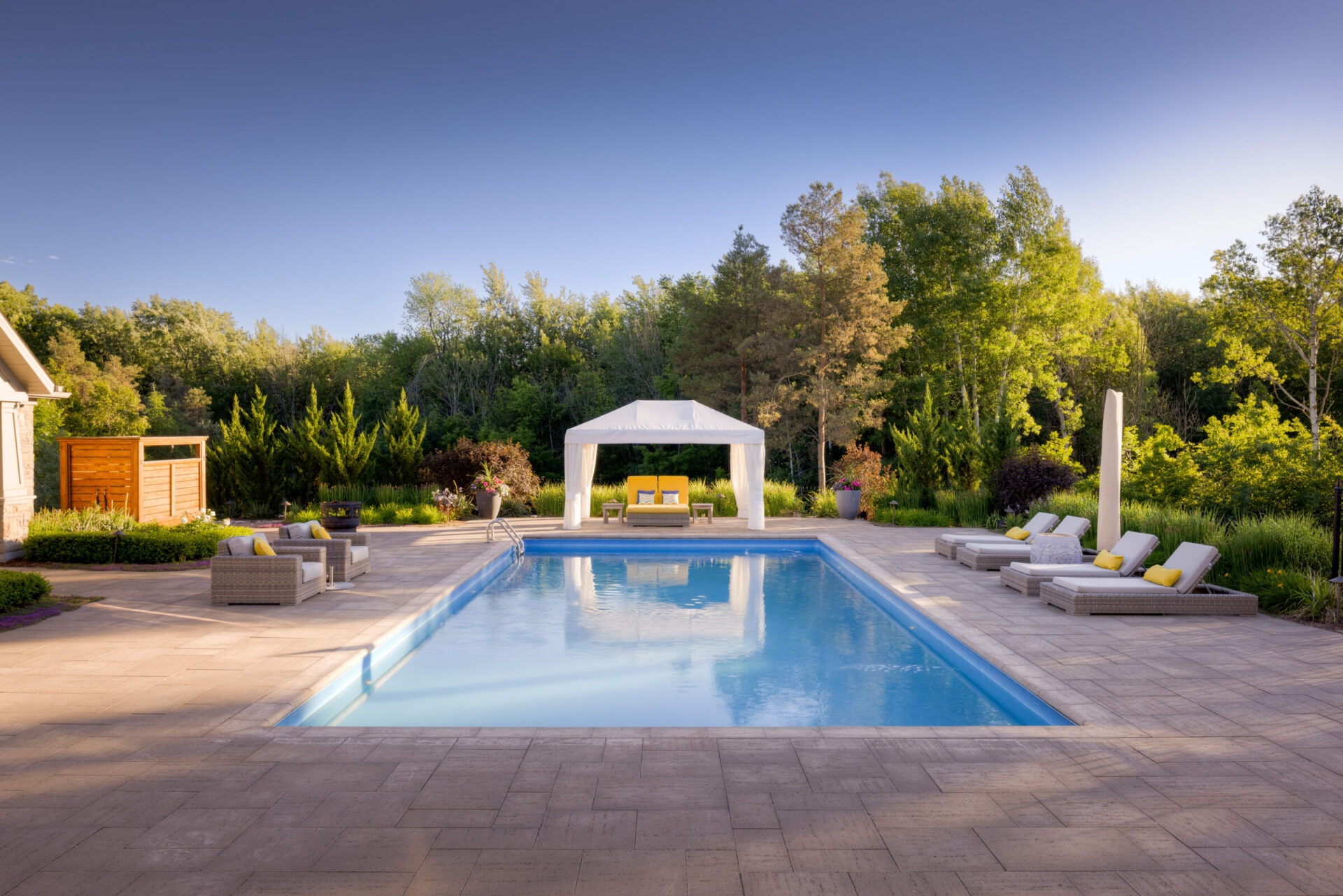An outdoor swimming pool with loungers and a white canopy, surrounded by a patio and lush greenery, under a clear sky during the day.