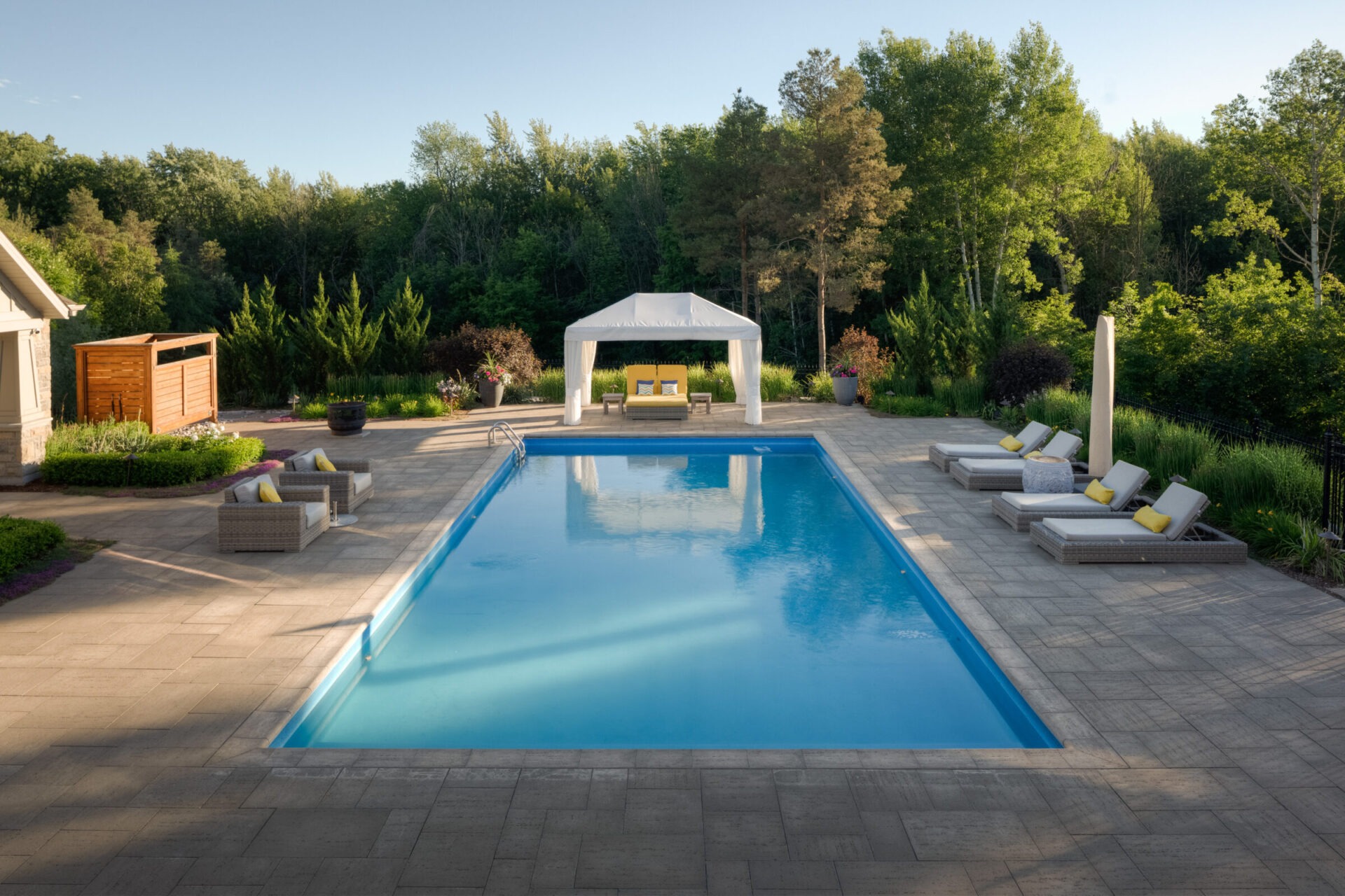 This image shows an outdoor residential swimming pool with chaise lounges and a gazebo, surrounded by trees, a garden, and a clear sky above.