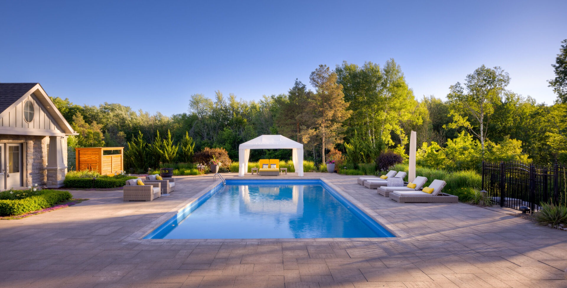 This image shows an elegant backyard with a large swimming pool, a canopy-covered seating area, lounge chairs, and lush greenery under a clear blue sky.