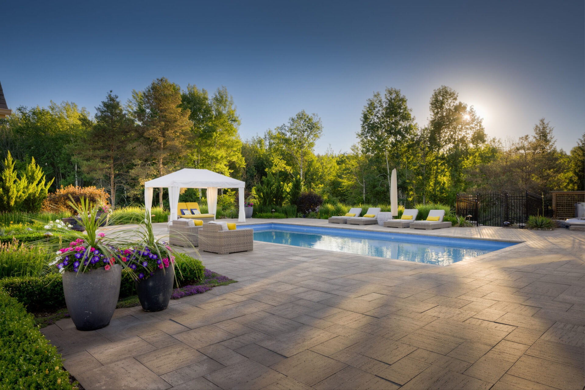 An outdoor pool area with loungers, a gazebo, and large planters surrounded by lush landscaping. The setting sun creates a tranquil atmosphere.