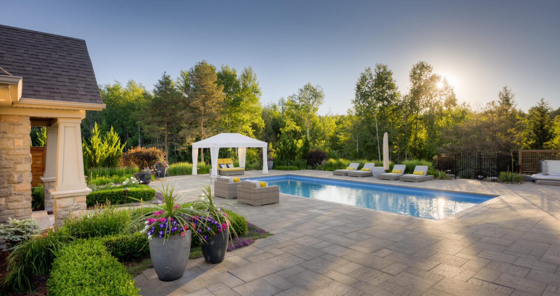 Luxurious backyard at sunset with a swimming pool, a gazebo, sun loungers, lush landscaping, and a clear sky, embodying tranquility and leisure.