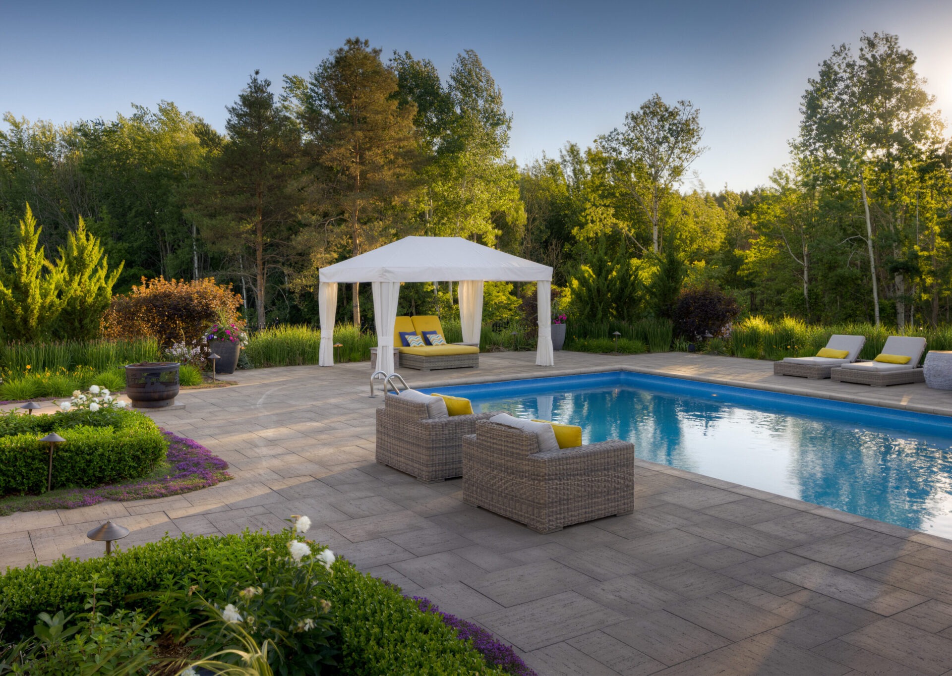 An outdoor pool with a white cabana and loungers surrounded by landscaped gardens, trees, and a clear sky as the sun sets.