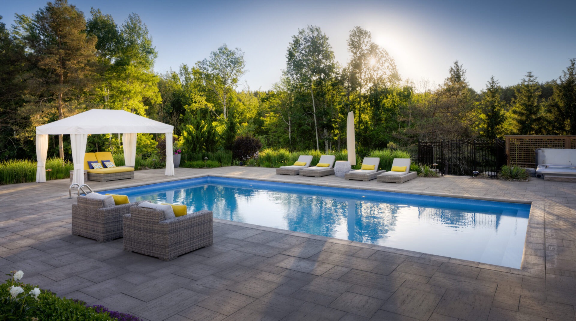 An elegant outdoor swimming pool with loungers and a gazebo, surrounded by lush greenery and trees, reflecting a serene, sunlit sky.