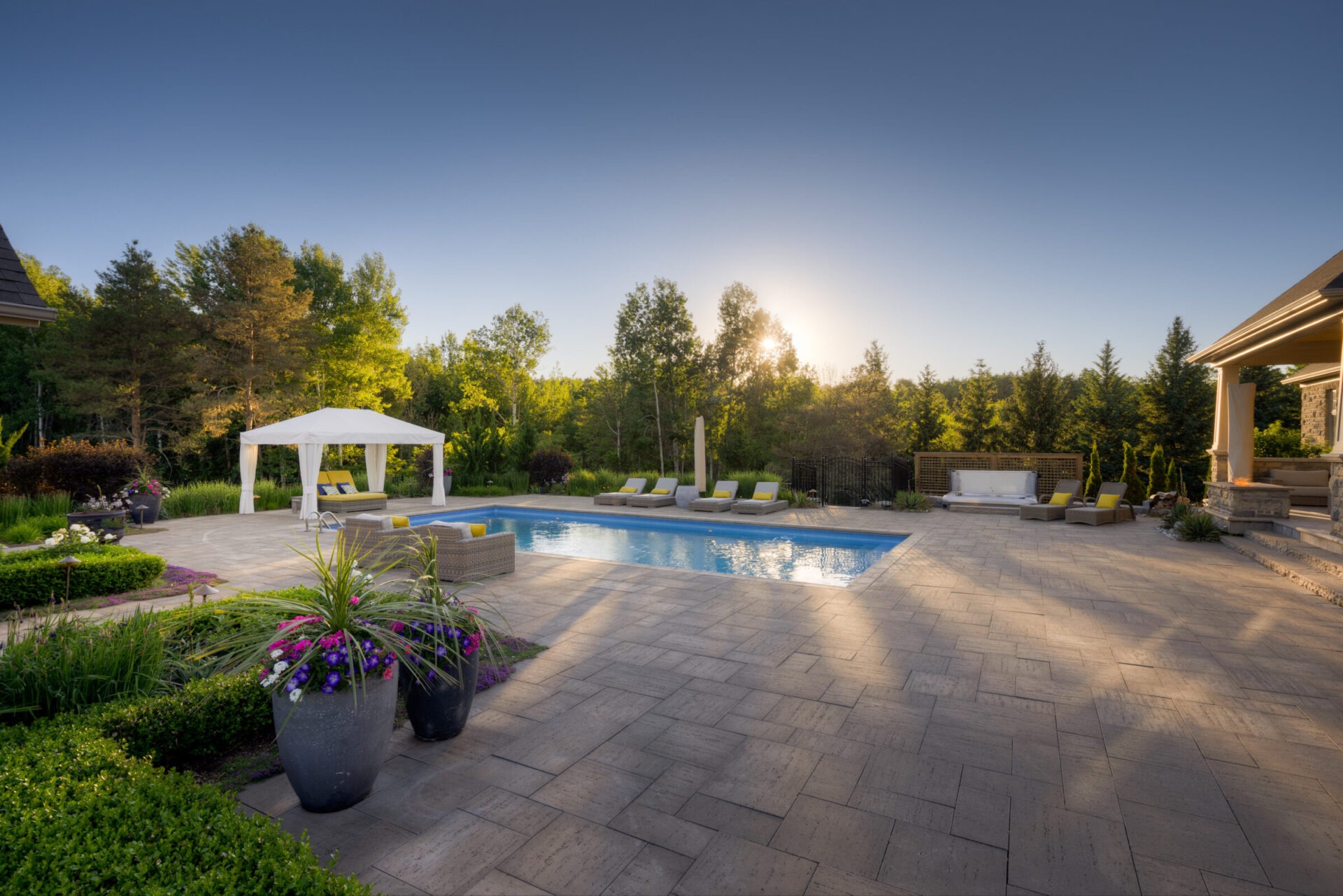 A luxurious backyard with a pool, sun loungers, a gazebo, tiled patio, and landscaped garden at sunset, surrounded by lush greenery and clear skies.