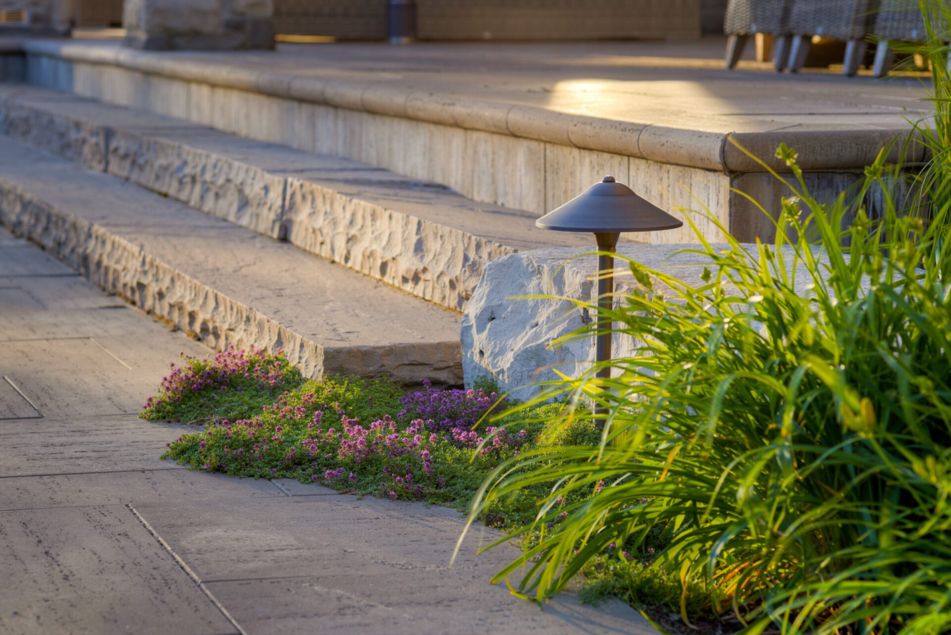 This image displays an outdoor setting with a pathway, a stone bench, ornamental grasses, and a metal lamp post, indicating a landscaped area, likely in a park or garden.