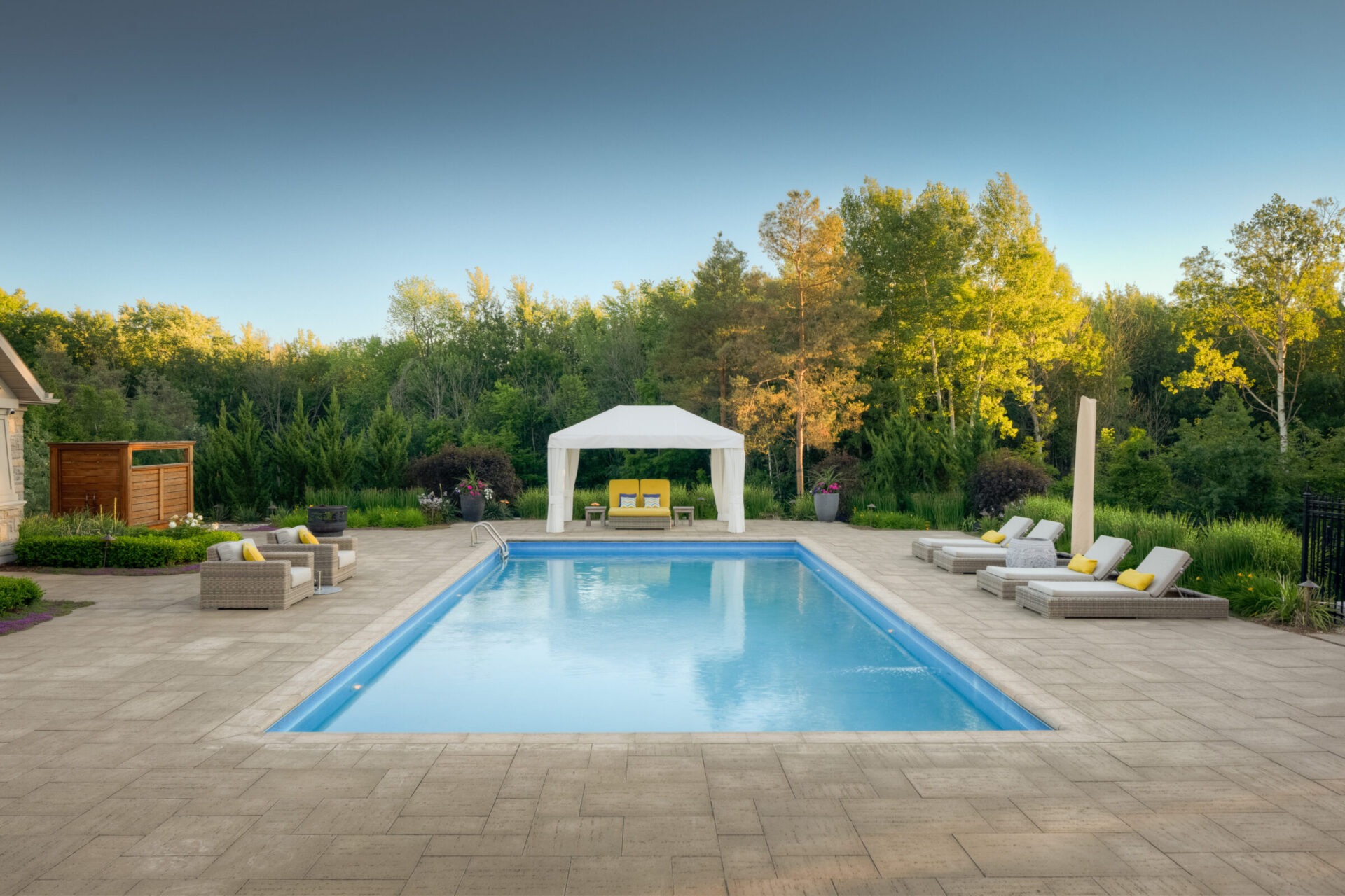 An outdoor residential pool with loungers, a white canopy, surrounded by landscaped garden and trees against a clear blue sky during daytime.