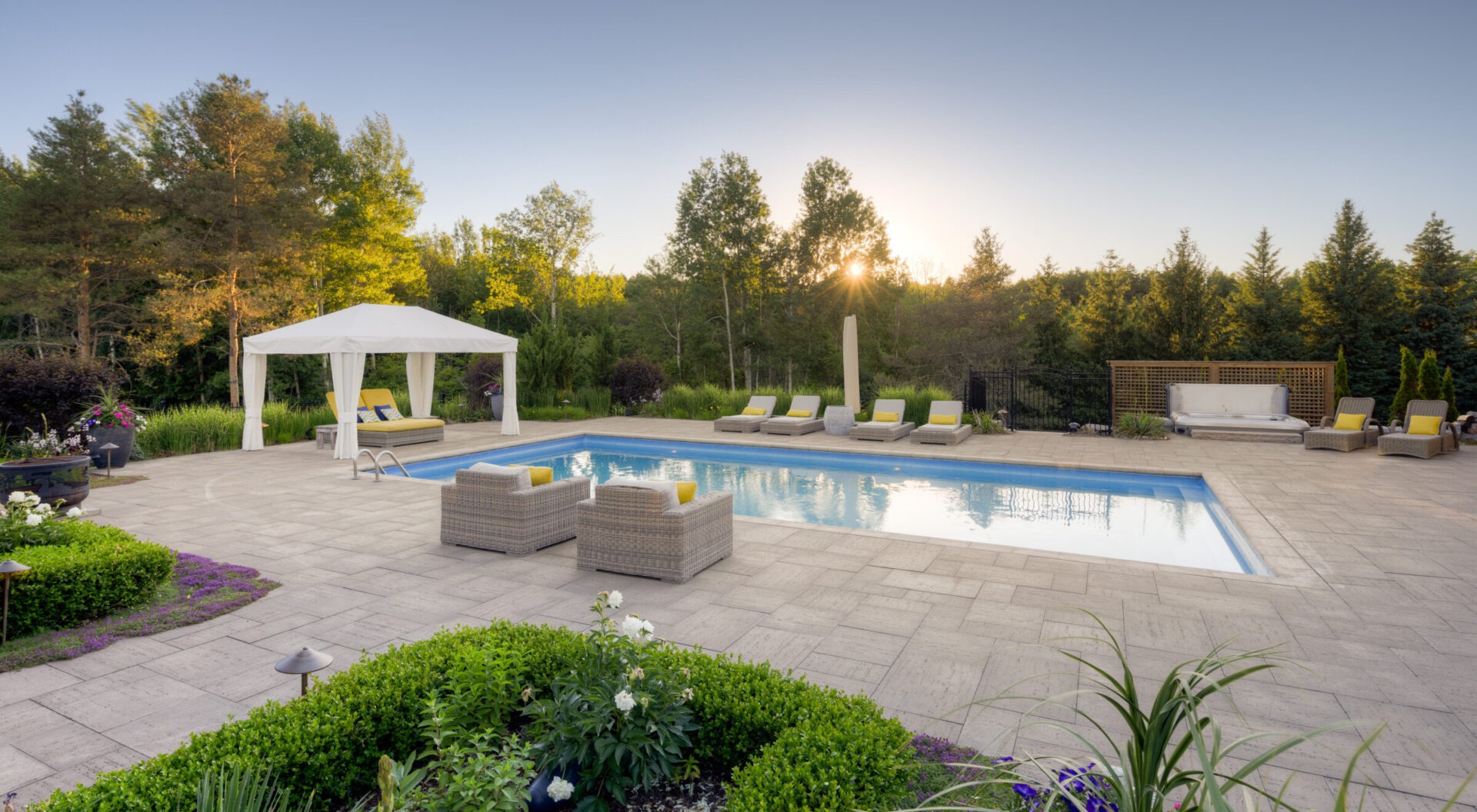 A luxurious backyard with a swimming pool during sunset, surrounded by trees, features a gazebo, loungers, and a seating area with plush furniture.