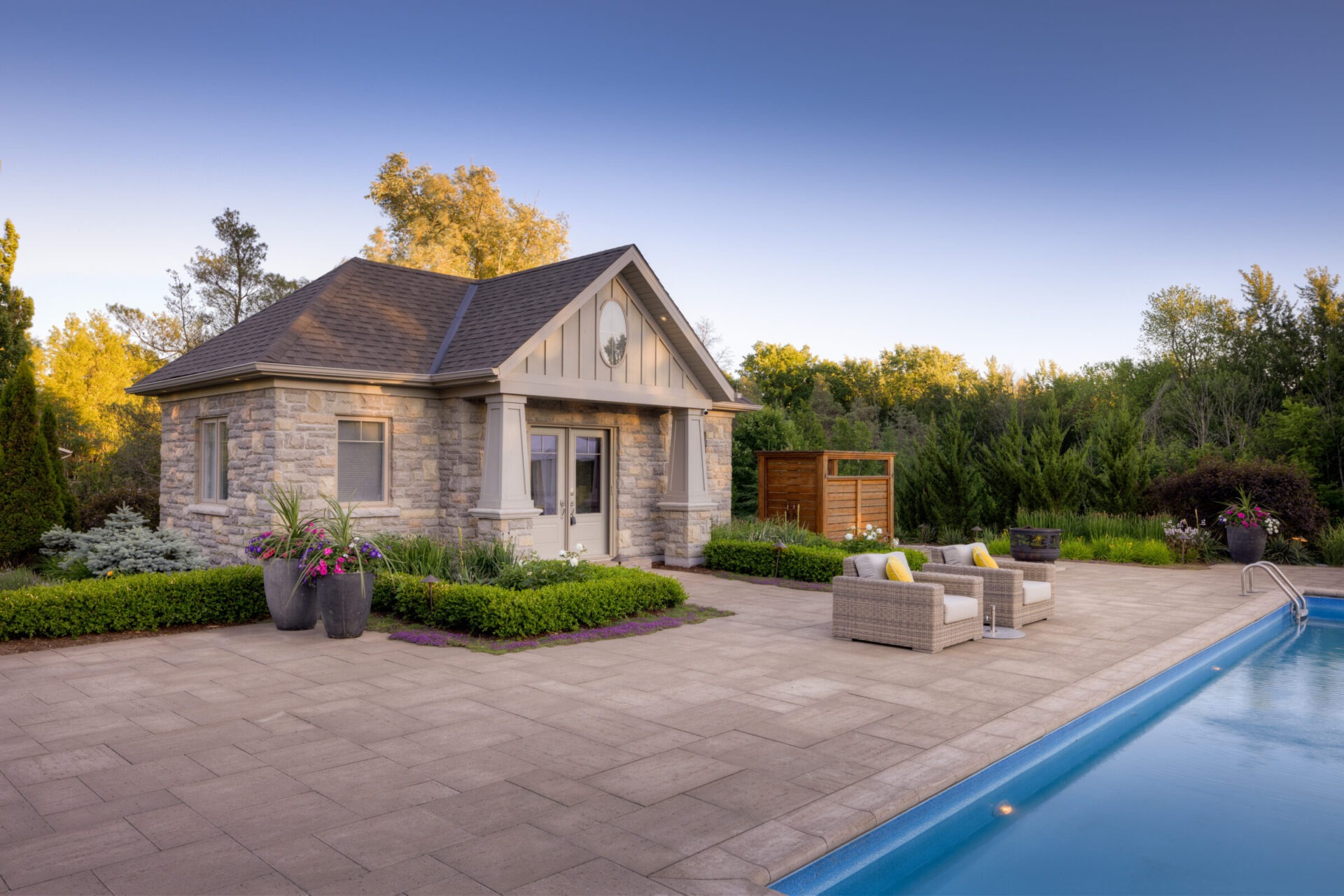 This is an image of a single-story stone house with a gabled roof, next to a swimming pool, outdoor furniture, and lush landscaping during twilight.