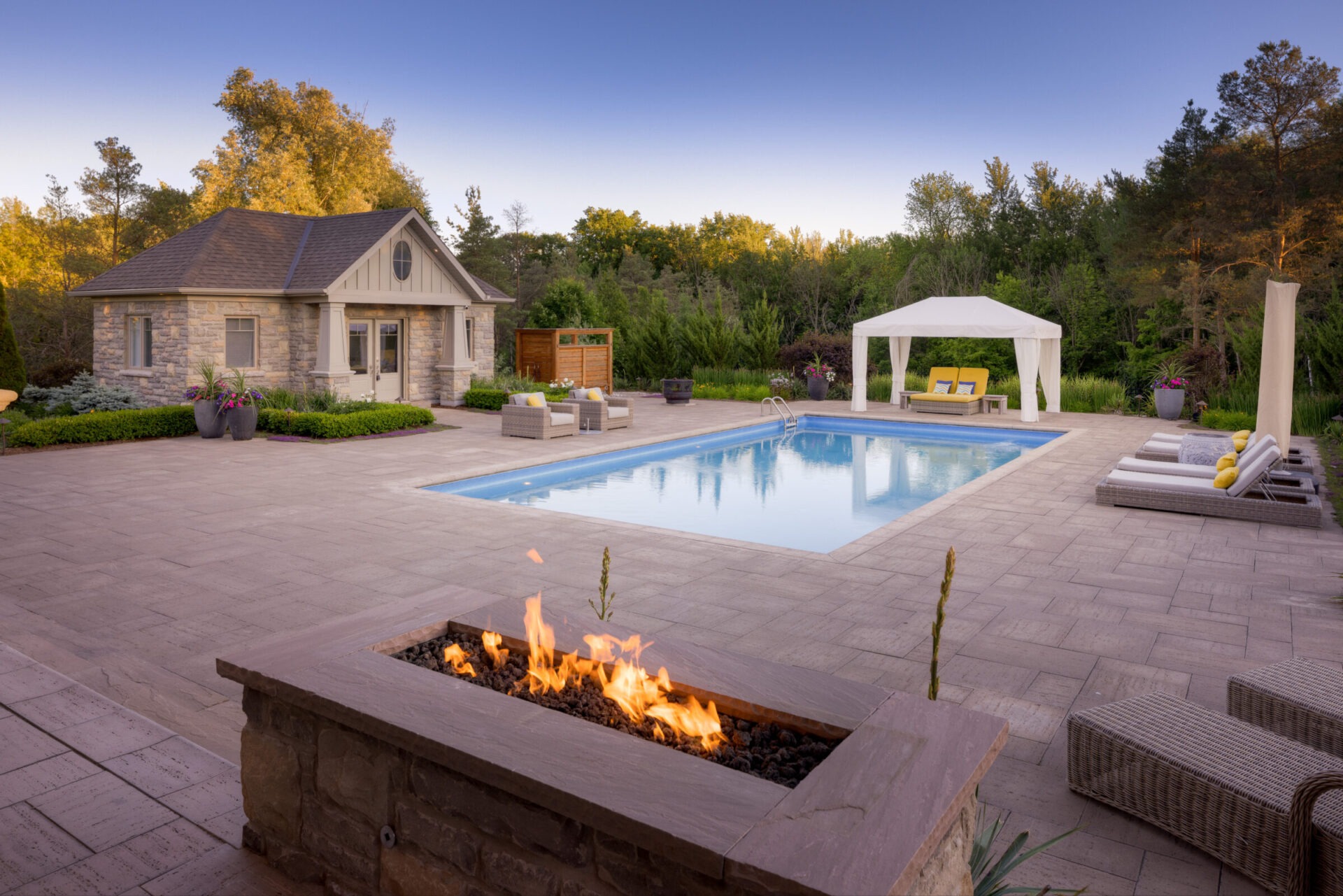 A luxurious backyard with a swimming pool, outdoor fire pit, patio furniture, a pergola, and a stone house surrounded by lush greenery during twilight.